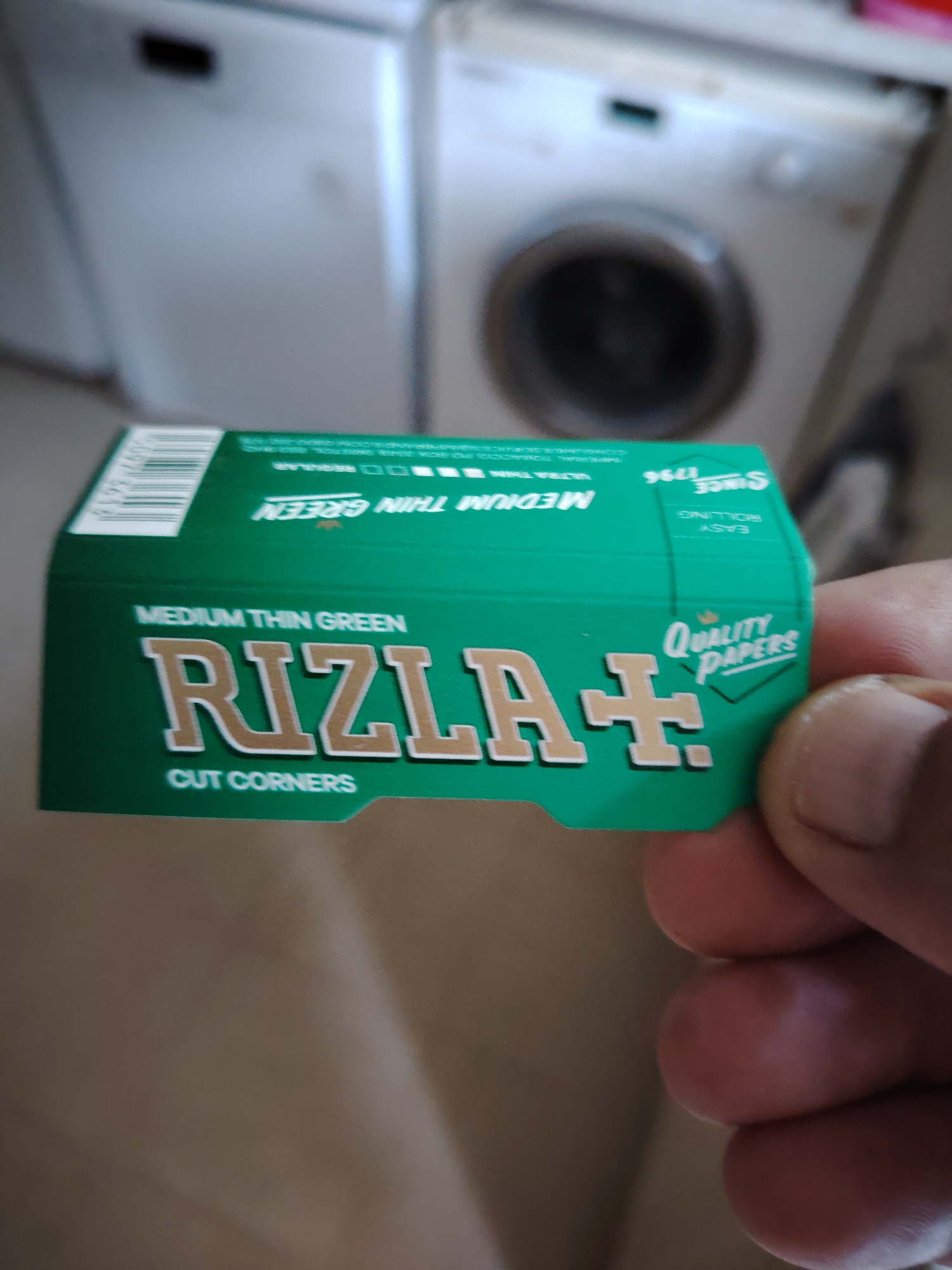 The image shows a close-up of a hand holding a pack of Rizla rolling papers. The pack is green with white and gold lettering. Text on the pack reads "MEDIUM THIN GREEN", "RIZLA+", "QUALITY PAPERS", and "CUT CORNERS". In the background, two washing machines are visible. The scene is indoors. There is no indication of the location in the city of Huntingdonshire, United Kingdom.