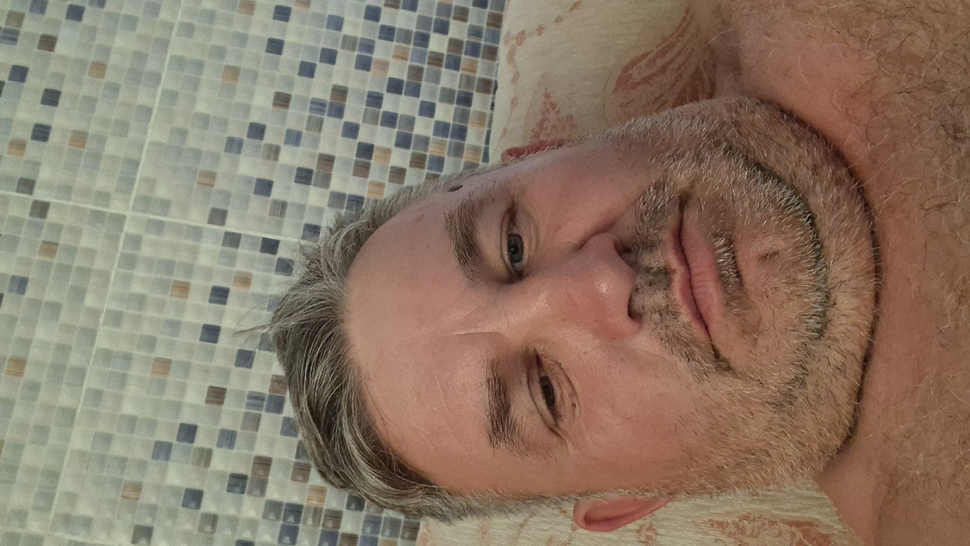 An over-the-shoulder selfie is taken from a low angle, looking up at a middle-aged man. He has graying hair, thinning on top, and a short beard. His eyes are partially closed, and his mouth is slightly open. The background is a tiled wall with a mosaic pattern of blue, gray, and orange squares. The man appears to be lying down, possibly in bed or on a sofa, as indicated by the soft fabric and pillow visible beneath him. The setting is indoors, likely a bathroom or bedroom, given the tiled wall. There is no indication of the specific location within Voinova, Moldova.