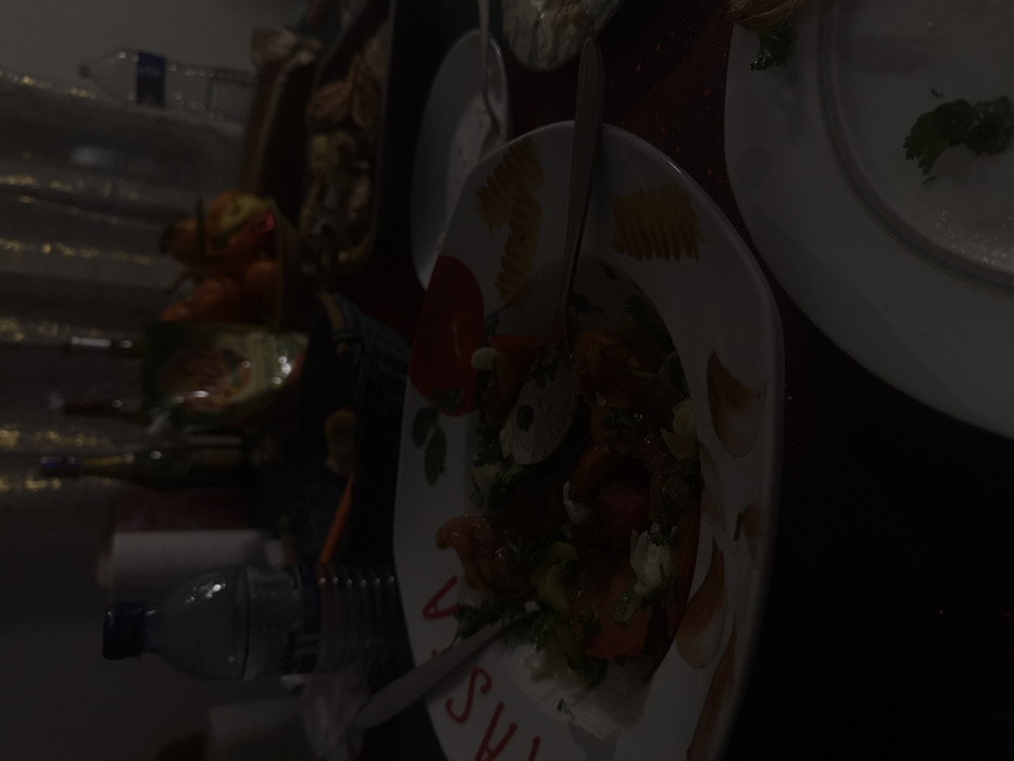 A dimly lit dining table features an arrangement of plates and food items. A salad with greens and tomatoes occupies a decorative plate with lettering. Beside it, a clean white plate contains a small green garnish. Further back, a bowl of oranges sits near bottles and glasses. Utensils are scattered amongst the dishes, suggesting a meal in progress. The composition is set against a dark backdrop.