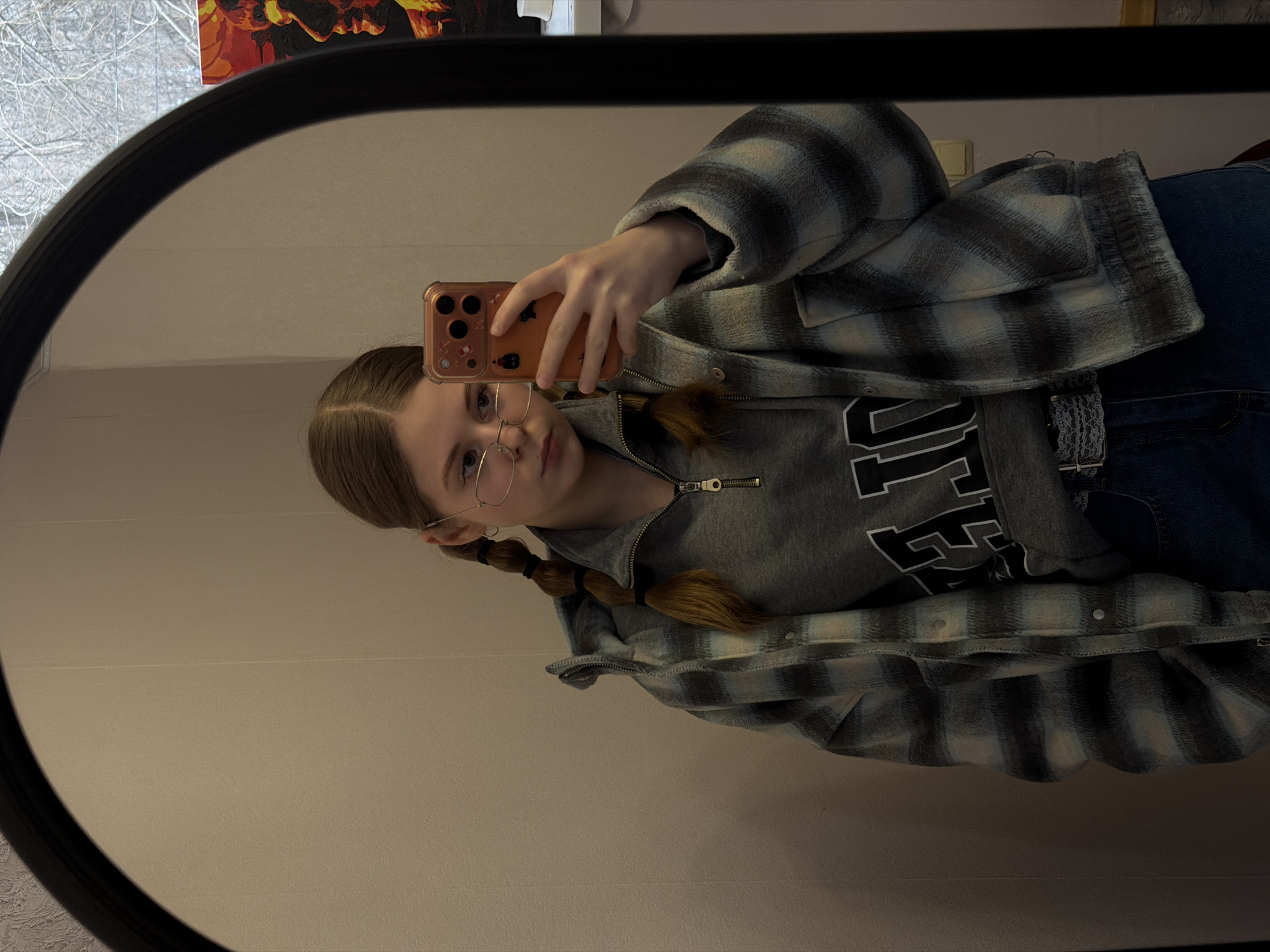 A young woman stands in front of a mirror, using a phone to capture a self-portrait. She is wearing glasses, and her hair is styled in braids. The woman has a grey hoodie with a black design and a plaid jacket, paired with blue jeans and a white lace belt. The setting is indoors, against a plain wall. The mirror is an oval-shaped object with a black frame. The reflections include the woman, the phone, and the surroundings. The location may be an apartment based on available visual clues. There is no forensic information to indicate a specific location in the city of Perm, Russia.