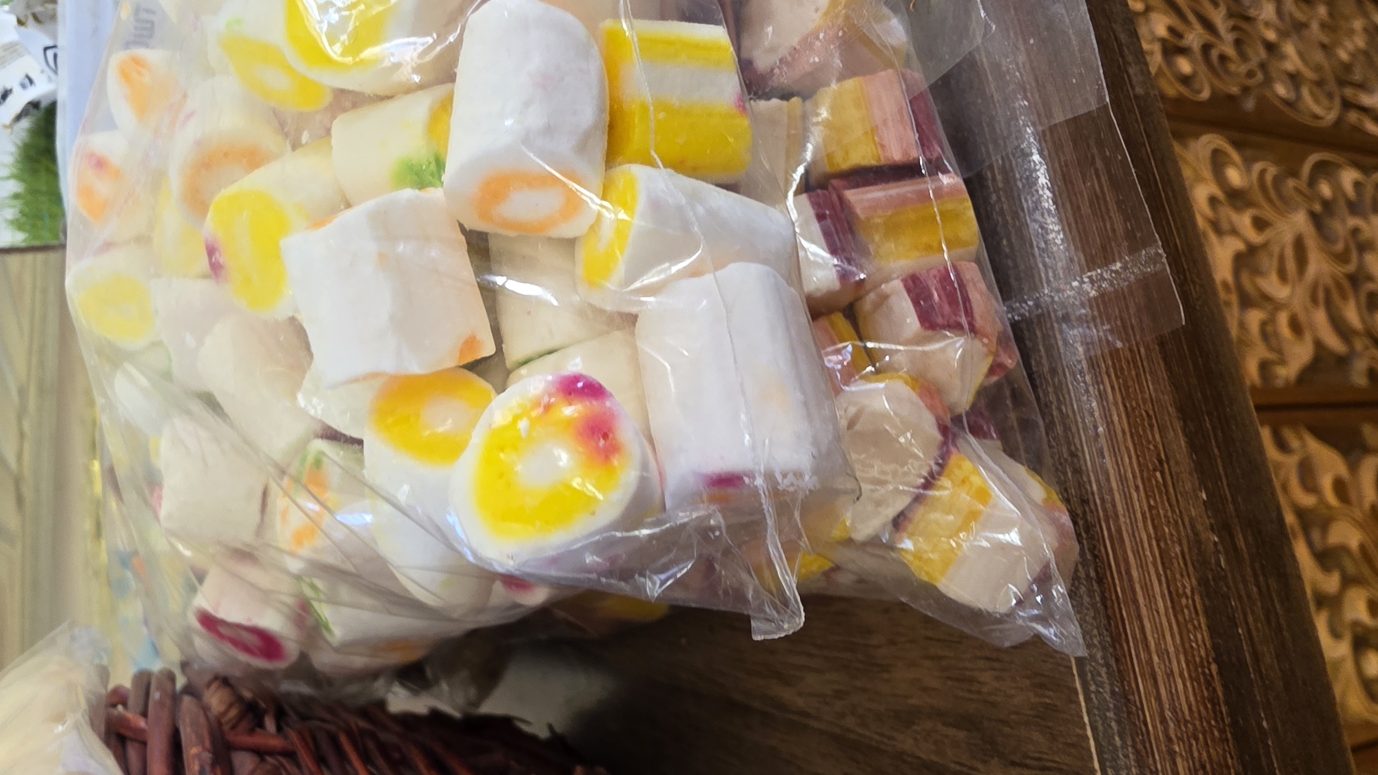 A close-up, eye-level shot shows a large plastic bag filled with individually wrapped, brightly colored hard candies. The candies are predominantly white with swirls of yellow, pink, orange, and green. Some candies are cylindrical with a layered, spiral pattern, while others are rectangular or cube-shaped with patches of color. The bag is placed on a wooden surface, possibly a table or shelf, with a woven basket visible in the upper left corner. The background is out of focus but appears to be an indoor setting with hints of green foliage and other indistinct objects. There is no indication of the specific location within Moscow, Russia.
