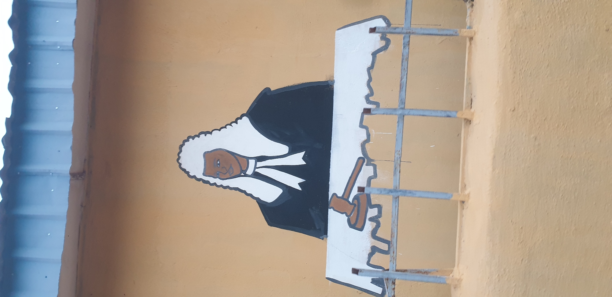 This is a medium-shot, eye-level, vertical, outdoor photograph of a mural painted on a light brown wall in Sabon Tasha, Nigeria. The mural depicts a person in judicial attire, featuring a white wig, a black robe, and a white jabot. The figure is seated behind a table covered with a white cloth, on which a brown gavel rests. The mural is positioned above a light grey metal railing. The background wall is textured and painted a light brown color. Above the wall, a corrugated metal roof with a blue-grey sheen is visible. The overall scene is brightly lit, suggesting daylight.
