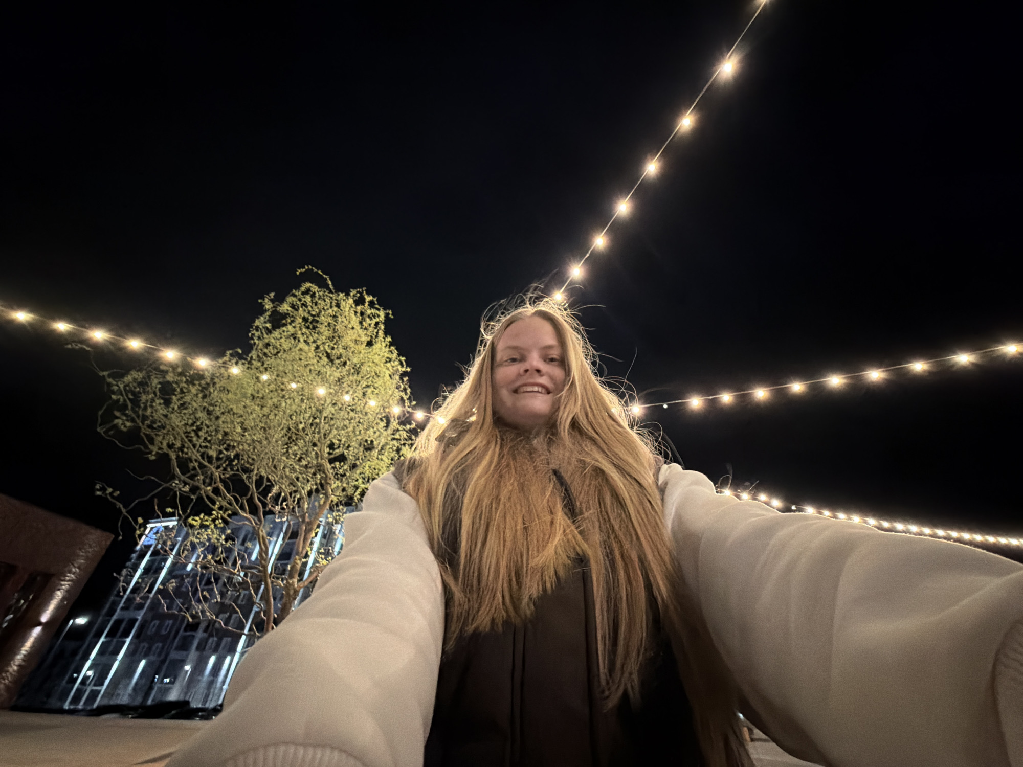 A young woman with long blonde hair is taking a selfie outdoors at night. She is wearing a light-colored sweatshirt with a dark vest. Behind her, there is a tree with string lights overhead. A building is visible in the background. It is not possible to confirm the location is Pervomaysky, Russia from the image.