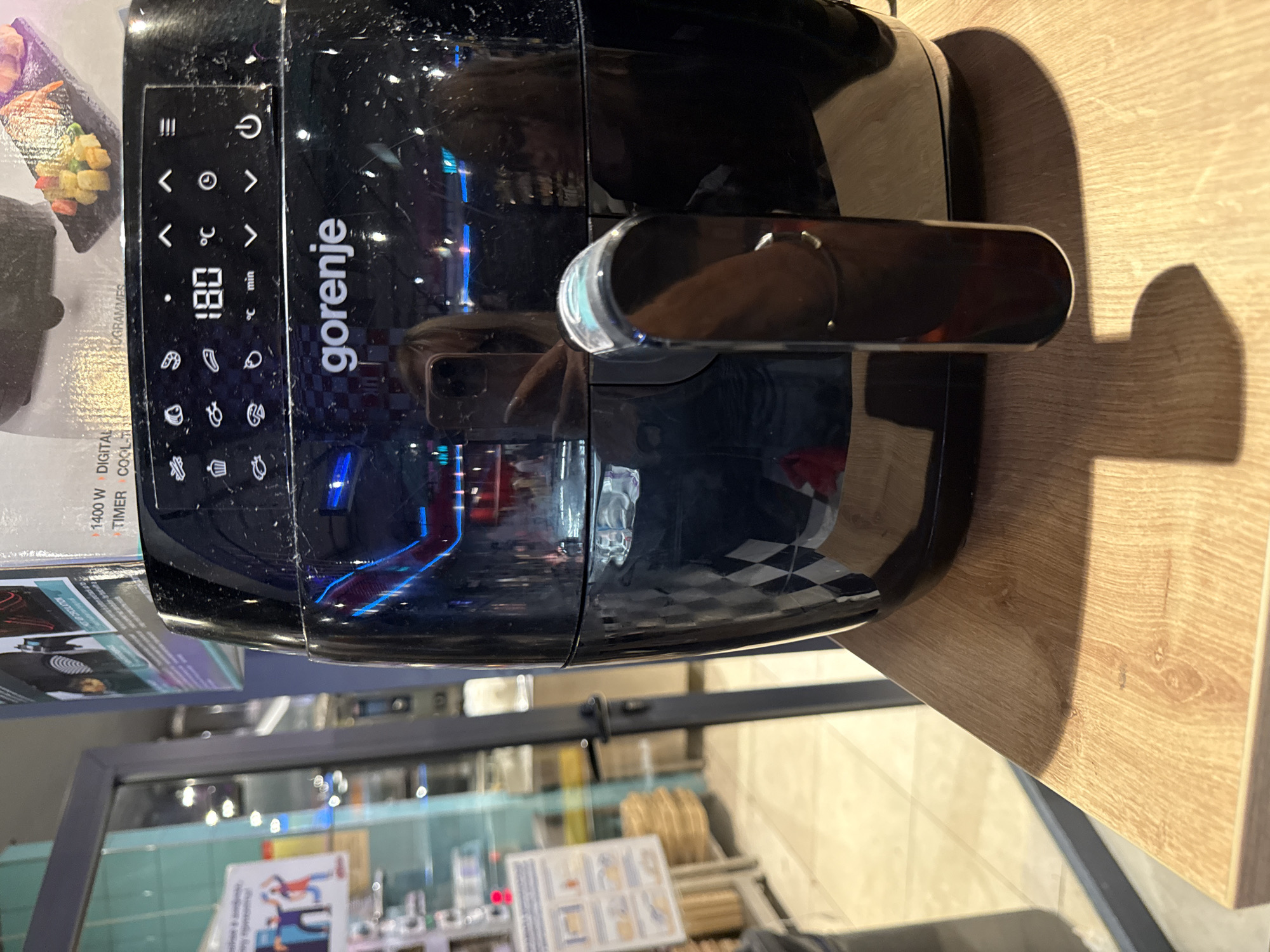 The image shows a black Gorenje air fryer placed on a wooden surface, possibly a countertop. The air fryer has a digital display showing "180 °C" and "min" for time. There are various icons and control buttons on the front panel, including a power button, temperature and time adjustment buttons, and pre-set cooking program icons. A chrome handle is attached to the front of the air fryer. The background is blurred, but appears to be an indoor setting with reflections of lights and possibly other appliances or products. Text visible on the air fryer's packaging or accompanying materials includes "1400 W DIGITAL PROGRAMMES" and "TIMER COOL-T". The overall lighting suggests it is daytime indoors. The scene is a product shot or a casual photo of a kitchen appliance.