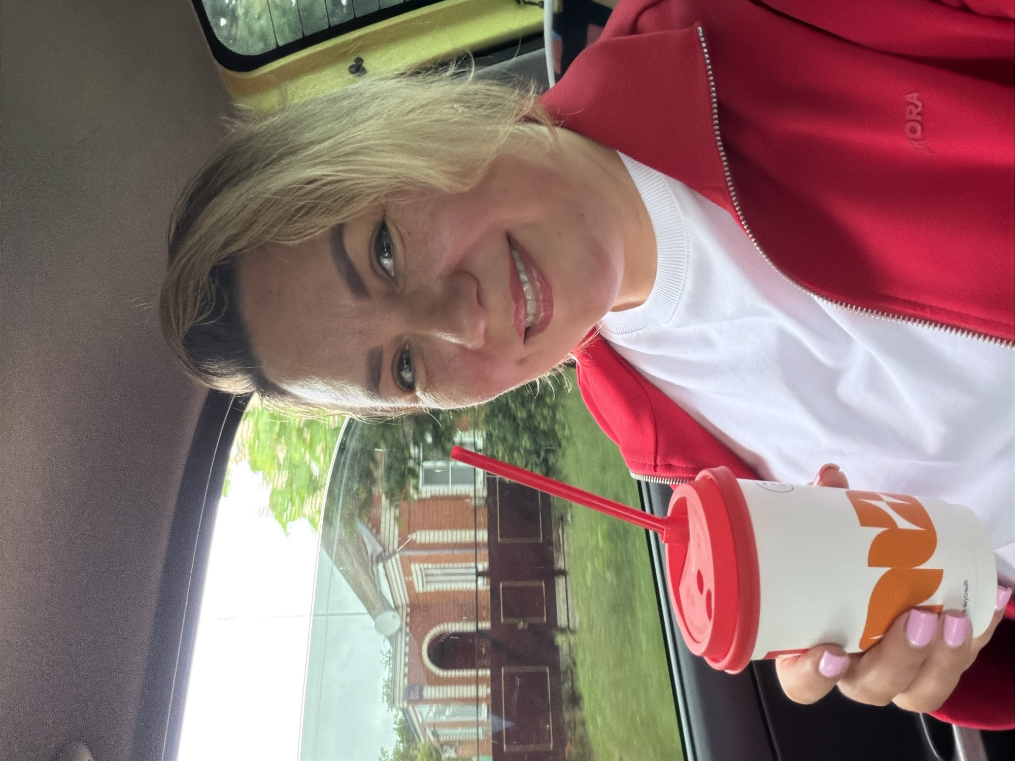 A woman in a red jacket and white shirt is inside a yellow car. She is holding a white and orange cup with a red lid and straw. The car window shows a reflection of a red brick building with arched doorways and windows. The building is surrounded by a green lawn. The location is likely Leningradskaya, Russia.