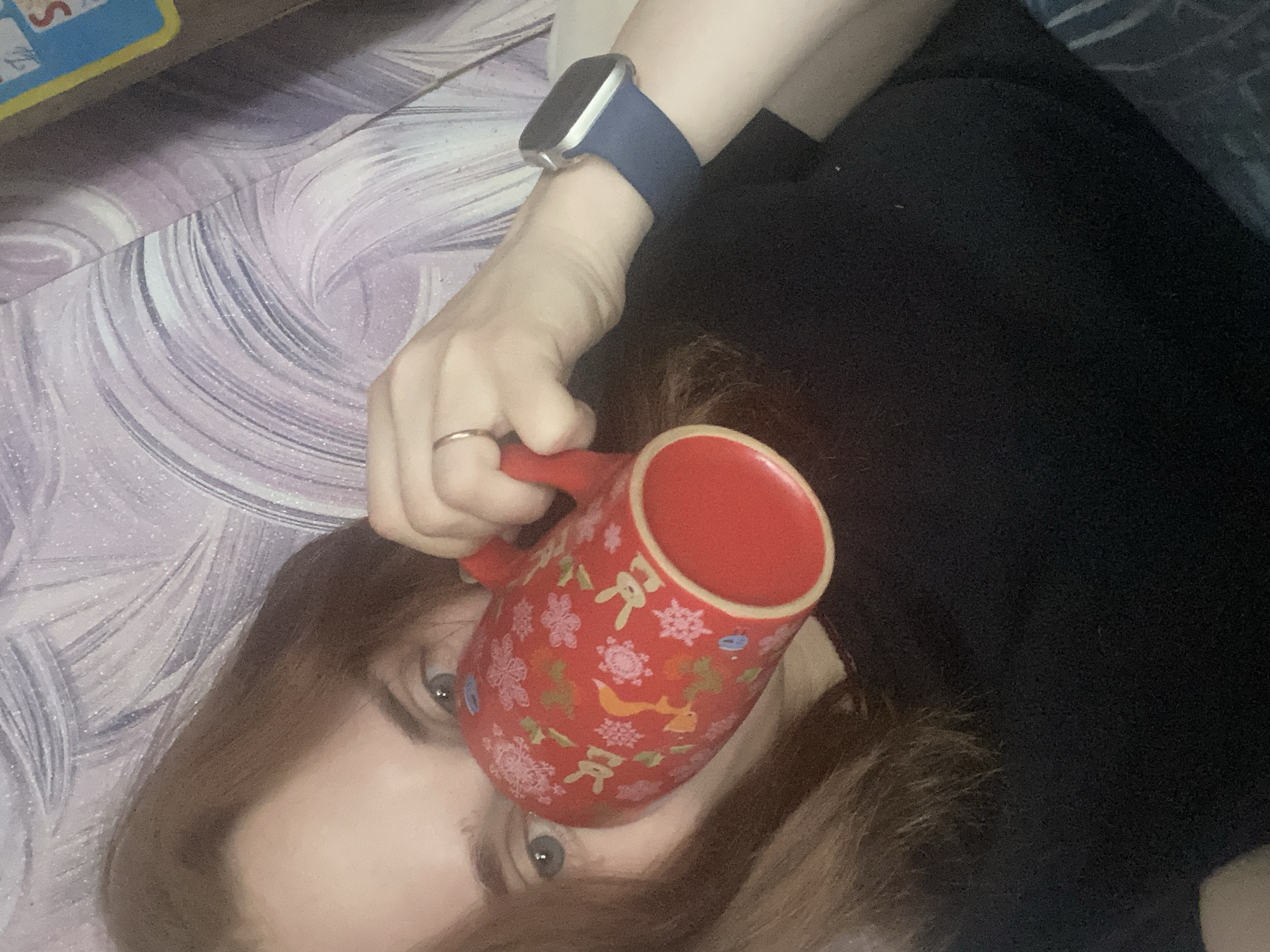 A person lies on a patterned surface, possibly a bed or blanket, holding a red mug with Christmas-themed designs. They are wearing a dark shirt and an Apple watch with a blue band. The location is not identified as Chaikovsky, Russia, from the given image.