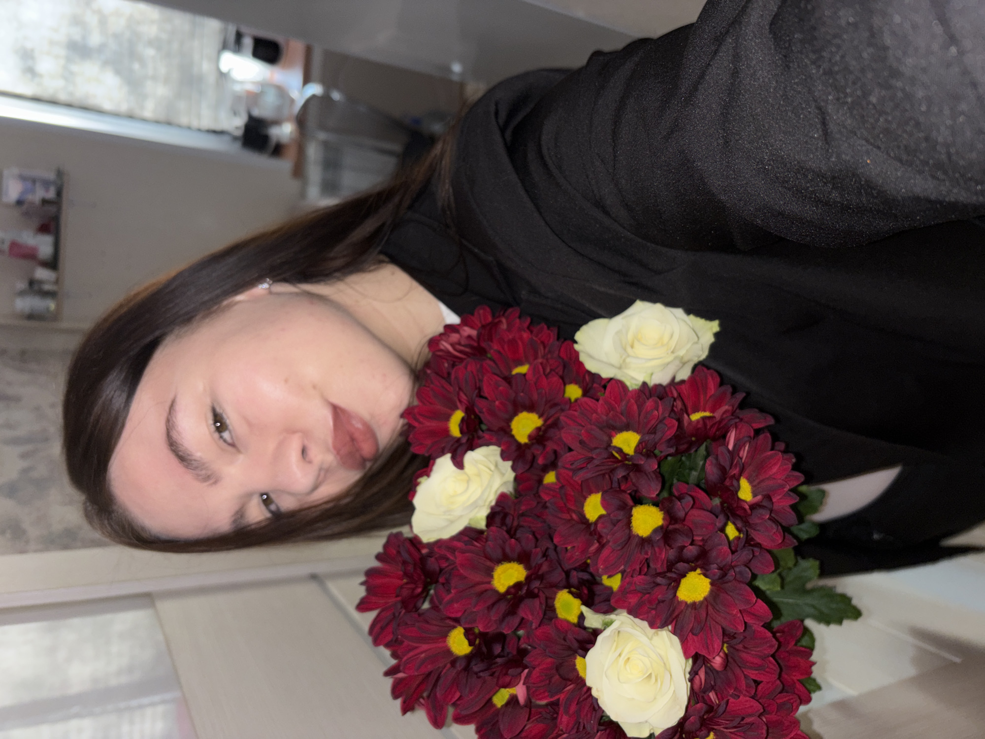 A young woman with brown hair is positioned in front of a bouquet of red and white flowers. She is wearing a black garment. The setting appears to be indoors, potentially in Perm, Russia, given the prompt's location reference. The camera angle is slightly tilted, showing a partial view of a room interior in the background. The image primarily focuses on the woman and the flowers.
