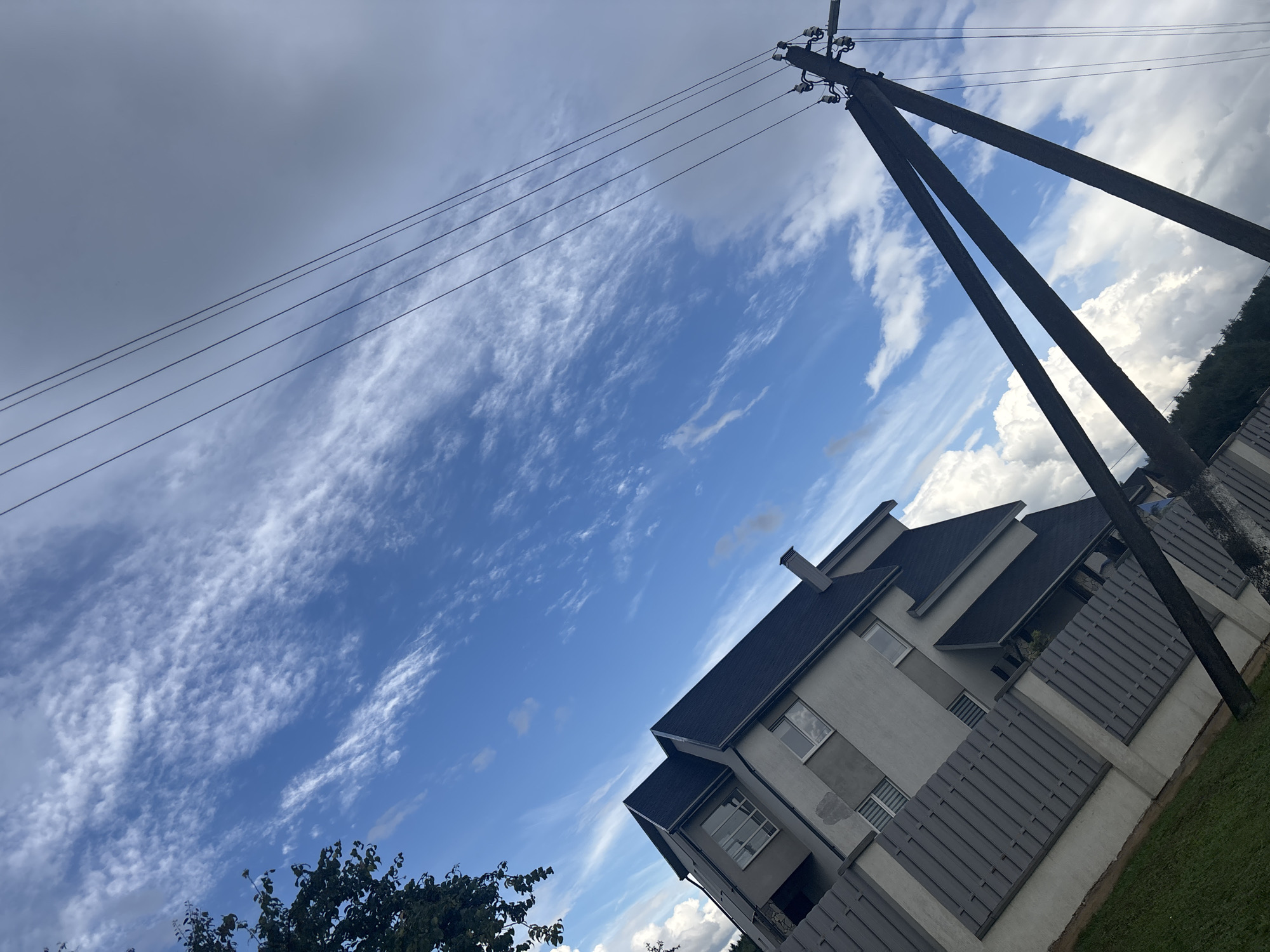A low-angle, outdoor, full shot captures a modern, grey, multi-story house with a dark grey roof. The house features several windows with white frames, and a grey slatted fence runs along its front. To the right of the house, a wooden utility pole with multiple crossbars and wires stands at an angle. The sky above is a vibrant blue, scattered with wispy white clouds and some larger, puffy white clouds closer to the horizon. A dark green tree branch is visible in the upper left corner. The scene appears to be set in a residential area, possibly in the Капыльскі сельскі Савет, Belarus, based on the provided location context. The lighting suggests it is daytime, with the sun casting shadows and illuminating the clouds.