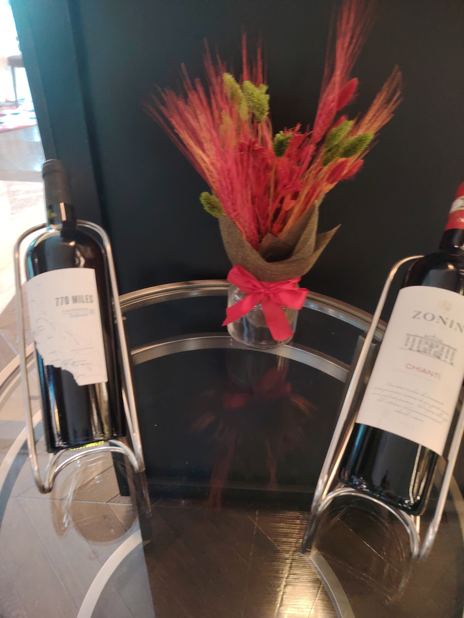 The image shows two bottles of red wine on a glass table, with a small arrangement of dried red and green decorative elements in the background. The bottle on the left has a white label that reads "770 MILES California". The bottle on the right has a white label that reads "ZONIN CHIANTI". A red ribbon is tied in a bow on a small glass vase. The lighting suggests an indoor setting, possibly a home or a commercial establishment. The image is not providing any information about the city of Konigil, Uzbekistan.