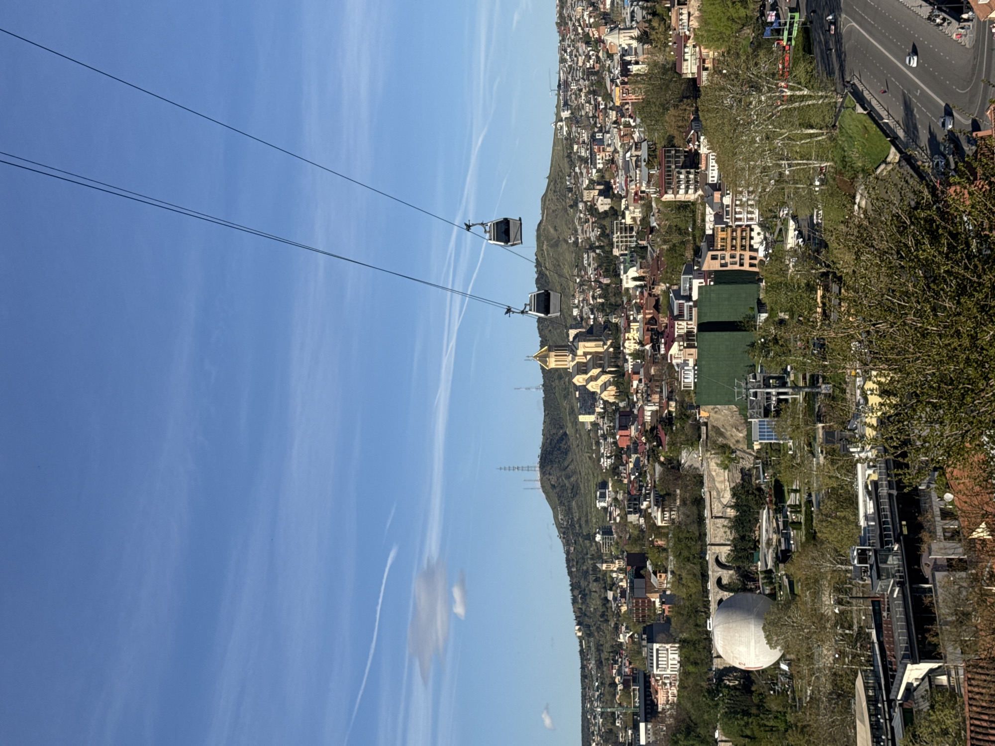The image depicts a view of the city of Tbilisi, Georgia, with two cable cars suspended against a blue sky with wispy clouds. The city sprawls along a hillside, featuring a mix of residential and commercial buildings with varying architectural styles. A road runs along the top of the hillside, with cars visible. Trees are interspersed throughout the city, adding patches of green. In the foreground, there's more vegetation, partially obscuring the view of some buildings. A large white dome-shaped structure is visible. In the distance, a mountain can be seen.