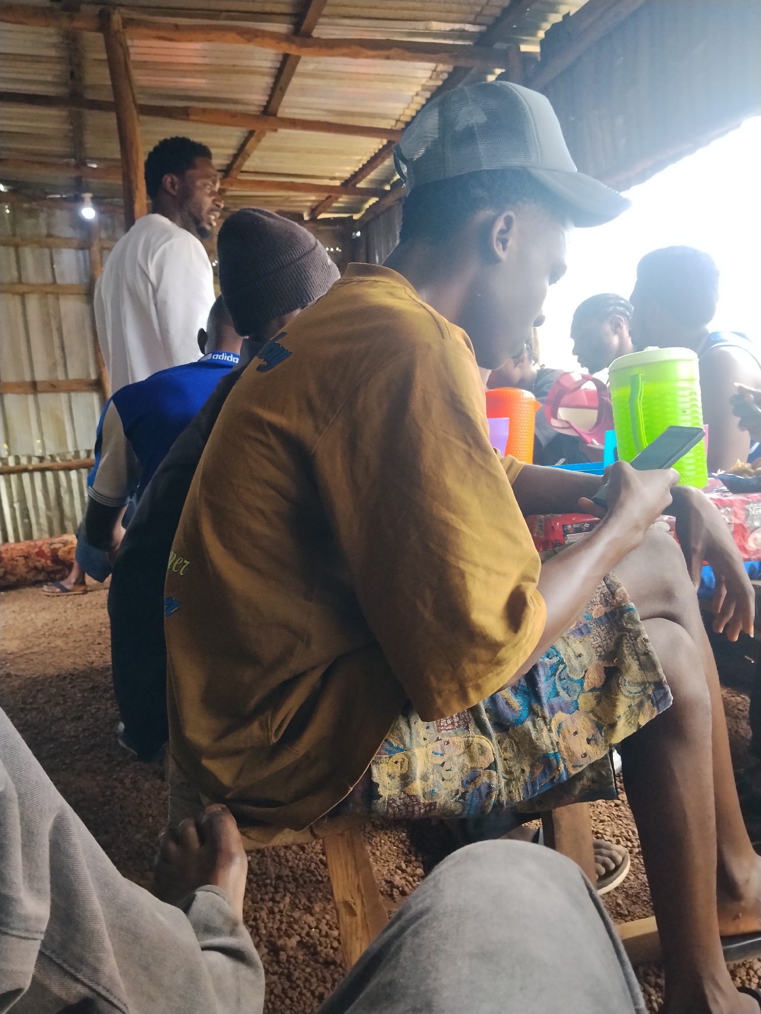 In Durumi, Nigeria, a group of young men are gathered under a rustic, open-air shelter with a corrugated metal roof supported by wooden beams. The ground is covered in what appears to be small pebbles or gravel. The men are seated or standing, engaged in various activities. In the foreground, one man, wearing a yellow t-shirt and a patterned skirt or wrap, is looking at his mobile phone. To his left, another young man is wearing a blue t-shirt with white and grey accents. Further back, a man in a white t-shirt is standing, facing away from the camera. Several colorful plastic cups and containers are visible on a table, along with what looks like a large, green, woven container. The lighting suggests it is daytime, with bright light coming from the right side of the frame, possibly indicating the sun.