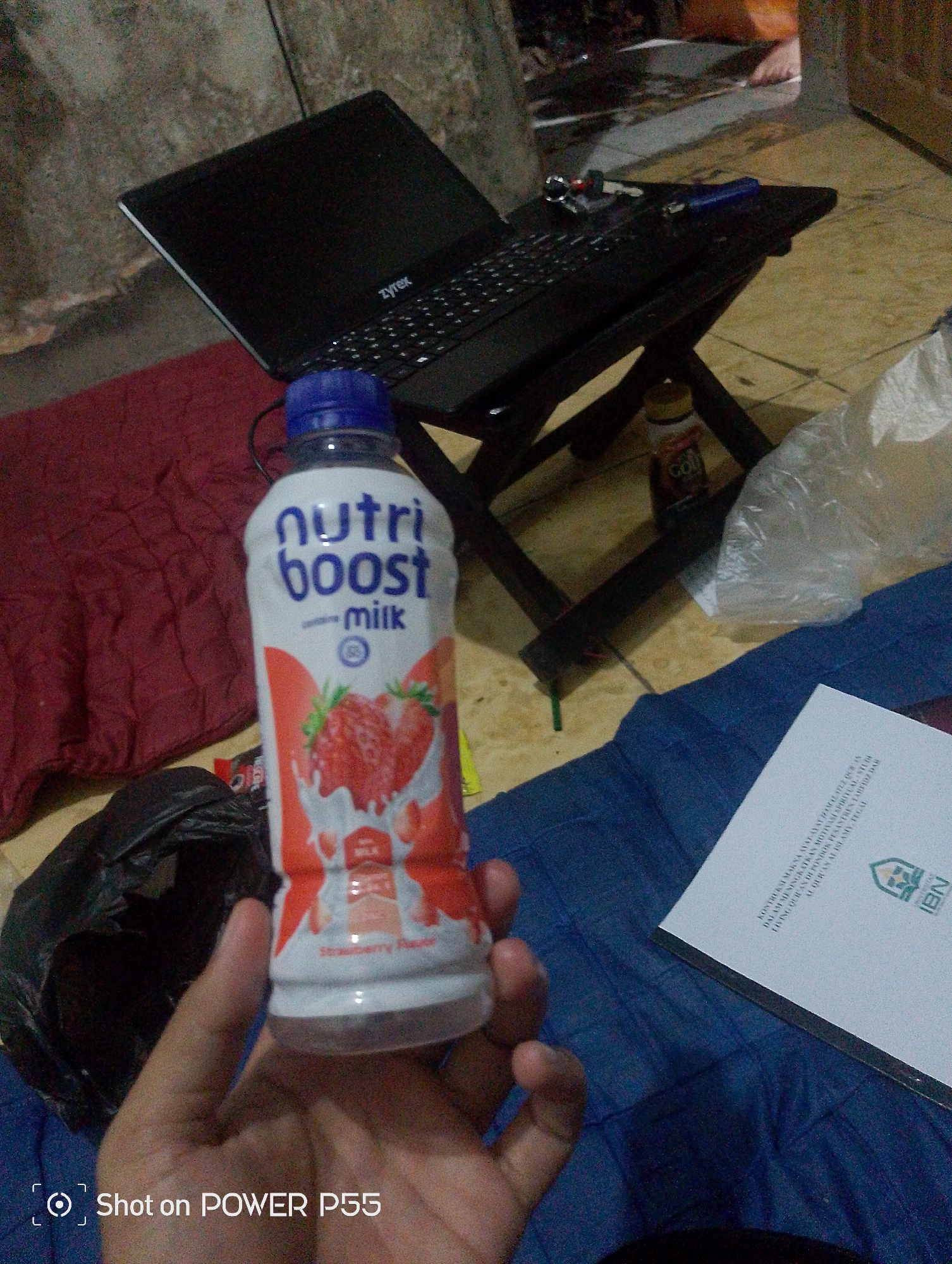 In Lebaksiu, Indonesia, a close-up shot focuses on a hand holding a bottle of NutriBoost strawberry milk. The hand is positioned in the foreground, partially obscuring a black plastic bag. Behind the bottle, a red blanket is visible. Further back, a black laptop rests on a dark wooden stand. To the right of the stand, a clear plastic bag contains a yellow-capped bottle. In the background, a light-colored wall with a textured surface is present. The image also includes a blue cloth spread out, with a white document featuring text and a logo on top.