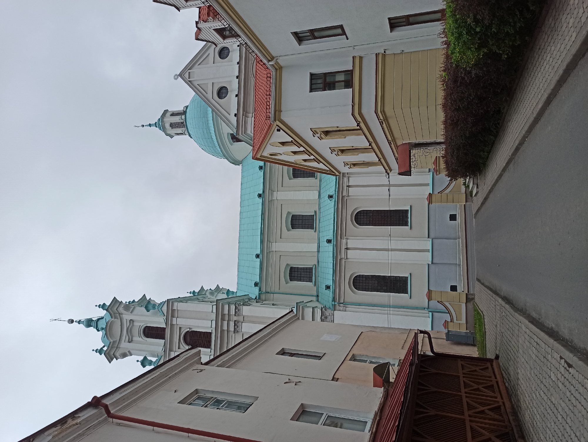 The image shows a street view in Hrodna, Belarus, featuring the facade of the Church of the Discovery of the Holy Cross and Bernardine Monastery. The church displays a prominent blue dome and spire, adjacent to buildings with varied architectural details, including white walls, red roofs, and multiple windows. A narrow street runs alongside the buildings, with a sidewalk paved with grey bricks and a road of dark asphalt. A hedge is visible alongside the buildings, adding some greenery to the urban scene. The sky is overcast. The photograph is taken from a lower angle looking upwards towards the buildings, giving emphasis on their vertical structures.