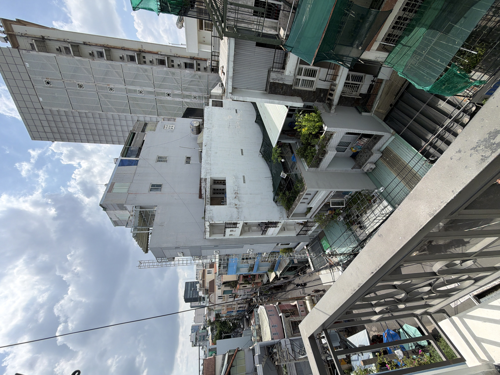 This image presents a panoramic view of a densely packed urban environment, likely captured from an elevated position such as a balcony or rooftop. The scene depicts a lively cityscape characteristic of Thủ Đức, Vietnam, under a partly cloudy sky during daytime.
In the immediate foreground, to the right, a dark gray metal railing with decorative circular cut-outs is visible, establishing the viewer's vantage point. Beyond this railing, a complex tapestry of multi-story buildings extends into the distance. These structures vary in height, age, and architectural style, ranging from narrow, tall residential houses to a more modern, larger building on the left with a grid-patterned facade. Many buildings feature balconies, some adorned with potted plants and greenery, while others display laundry hanging out to dry. Air conditioning units are visible on several exteriors.
Green protective netting and scaffolding are present on some buildings, hinting at ongoing construction or renovation. Rooftops are often equipped with water tanks. A narrow street or alleyway is visible below, where the blurred shapes of motorbikes and possibly pedestrians suggest daily activity, though no distinct individuals are clearly discernible. A dense network of overhead utility cables crisscrosses the view, a common sight in urban areas of Vietnam. The light suggests it is mid-day to afternoon. No explicit text is clearly visible within the frame.
