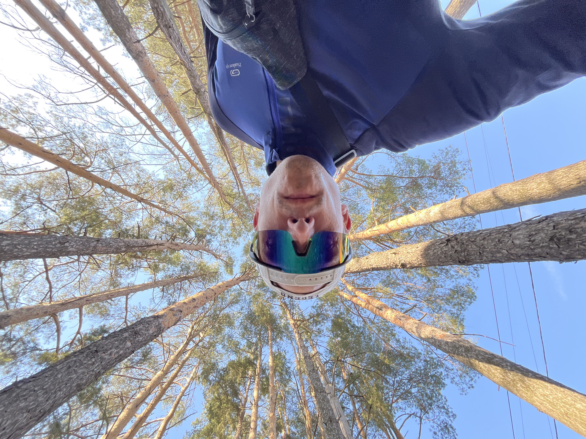 A low-angle shot looks upward through a forest of tall, straight pine trees toward a clear, light-blue sky. An adult male is in the foreground, the camera positioned directly below them, creating an inverted perspective of the person’s face and upper body. The man wears reflective sunglasses, a white baseball cap, and a blue and gray jacket. The trees’ trunks are a light-brown color and the branches are sparsely covered with green pine needles. The overall lighting is bright and appears to be taken during daytime. The location is the city of Chaikovsky, Russia, based on previously learned information.