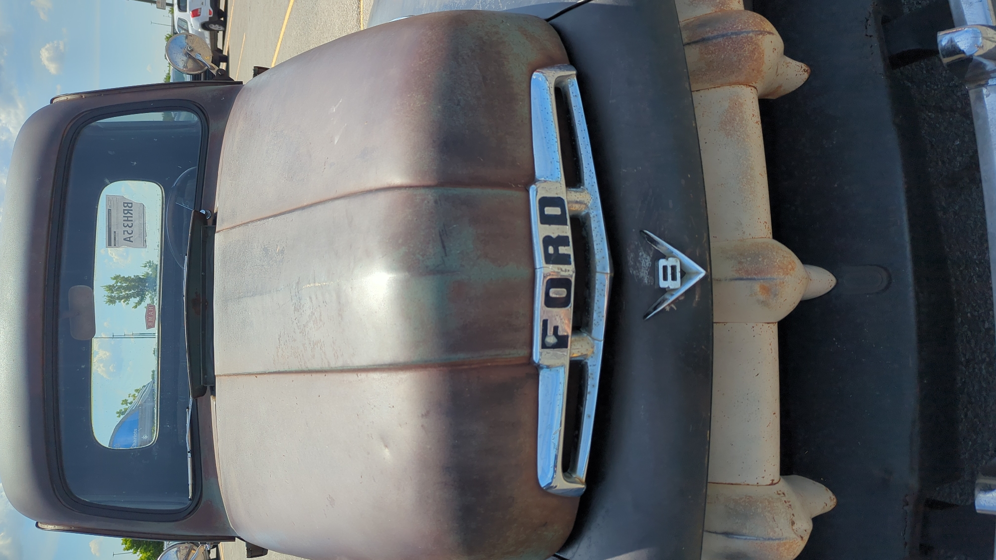 The image shows a close-up frontal view of a rusty, vintage Ford truck. The truck's hood, grille, and bumper are the primary focus. The grille features the prominent "FORD" lettering in black against a chrome surround. Below the grille, a "V8" emblem is visible. The bumper is a two-piece chrome or painted metal component with rounded protrusions. The overall condition of the truck suggests it is weathered and has been exposed to the elements.
The truck is parked outdoors, likely in a parking lot, as indicated by the paved ground with yellow lines and other vehicles visible in the background, including a white SUV and a blue trailer. The sky above is partly cloudy with a bright, sunny atmosphere. A reflection of trees and buildings can be seen on the truck's windshield.
There are no people visible in the image. The context of Laval, Canada, is not directly observable within the image itself, but this description pertains to the visual content presented.