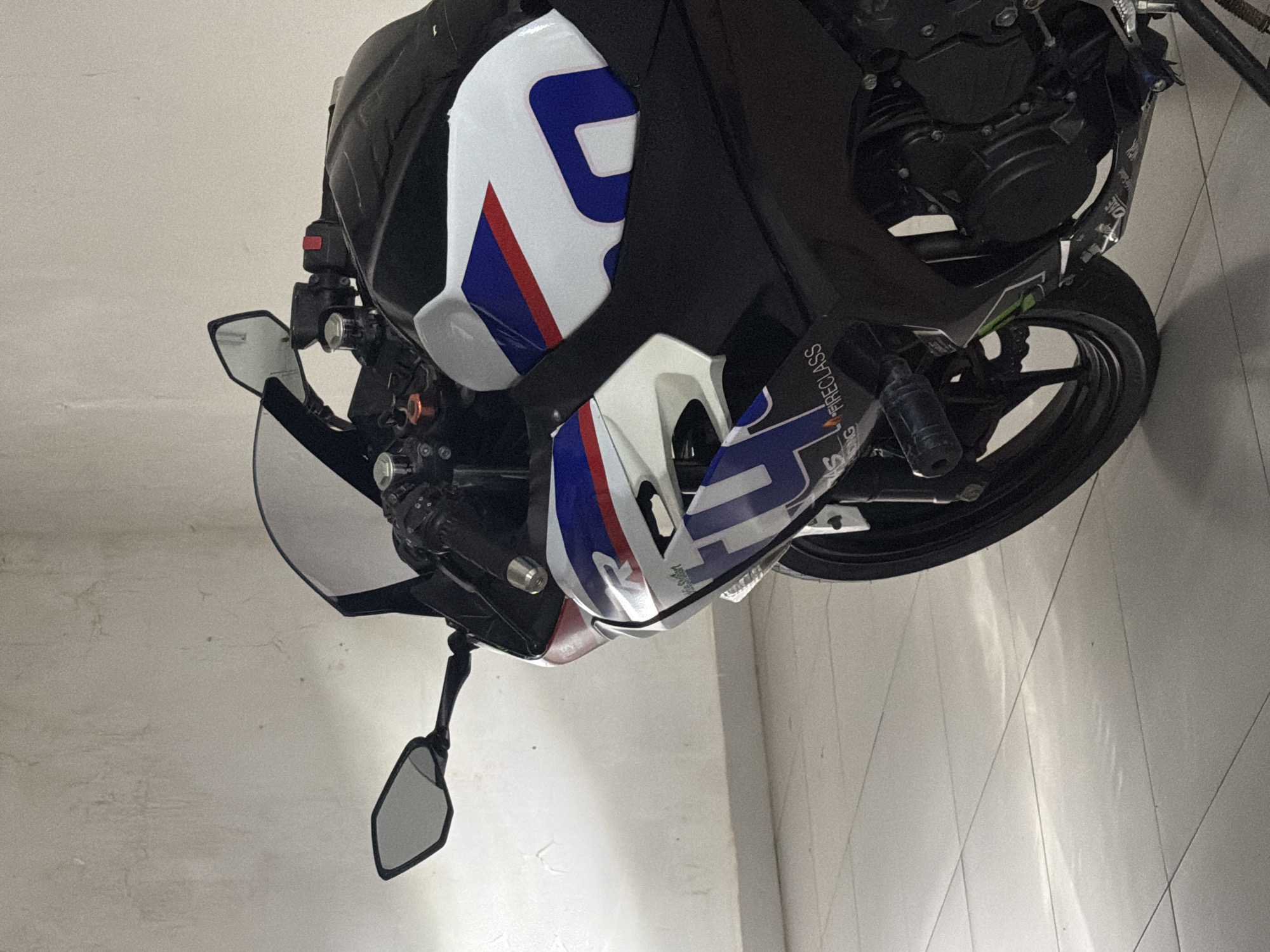 The image shows a motorcycle parked indoors against a white wall. The motorcycle is primarily black with white, blue, and red accents. Its mirrors are extended. The background is a simple white wall. There is no indication of the location being Đà Nẵng, Vietnam, or any specific city. There are no people visible in the image.