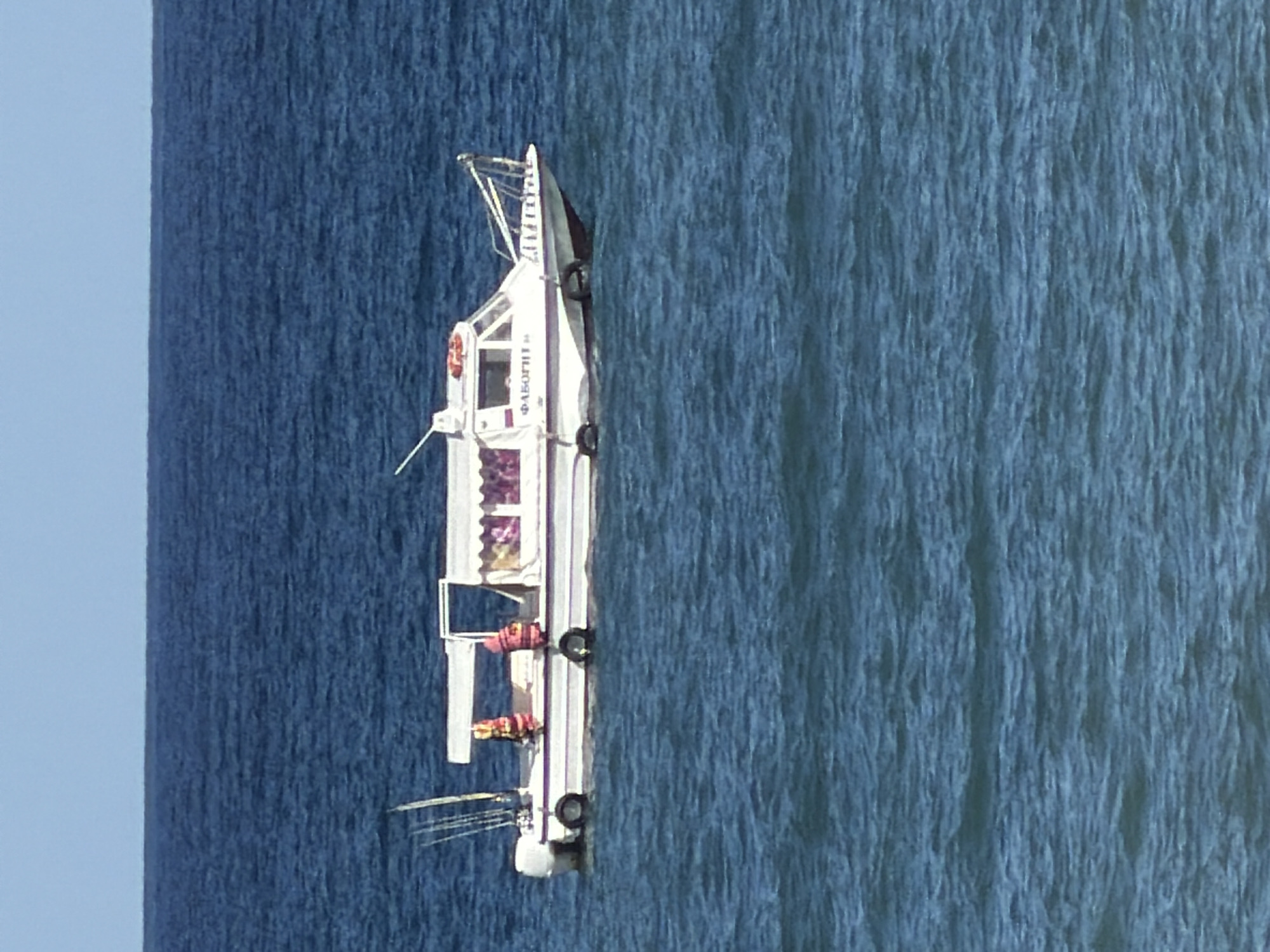 A white, multi-person boat with a white canopy is floating on the blue sea. The boat has a white hull with a dark blue stripe along the waterline. Two orange life vests are visible on the deck, one near the bow and one towards the stern. Fishing rods are mounted on the stern of the boat. There are two visible people on the boat, one wearing a red life vest and another wearing a similar vest. The windows of the boat's cabin are tinted. Text that reads "ФАБОПГН 8" is visible on the side of the cabin. The sea is calm with small waves. The sky is clear and blue. The scene is likely taking place in the city of Alushta, Ukraine, given the specific location.