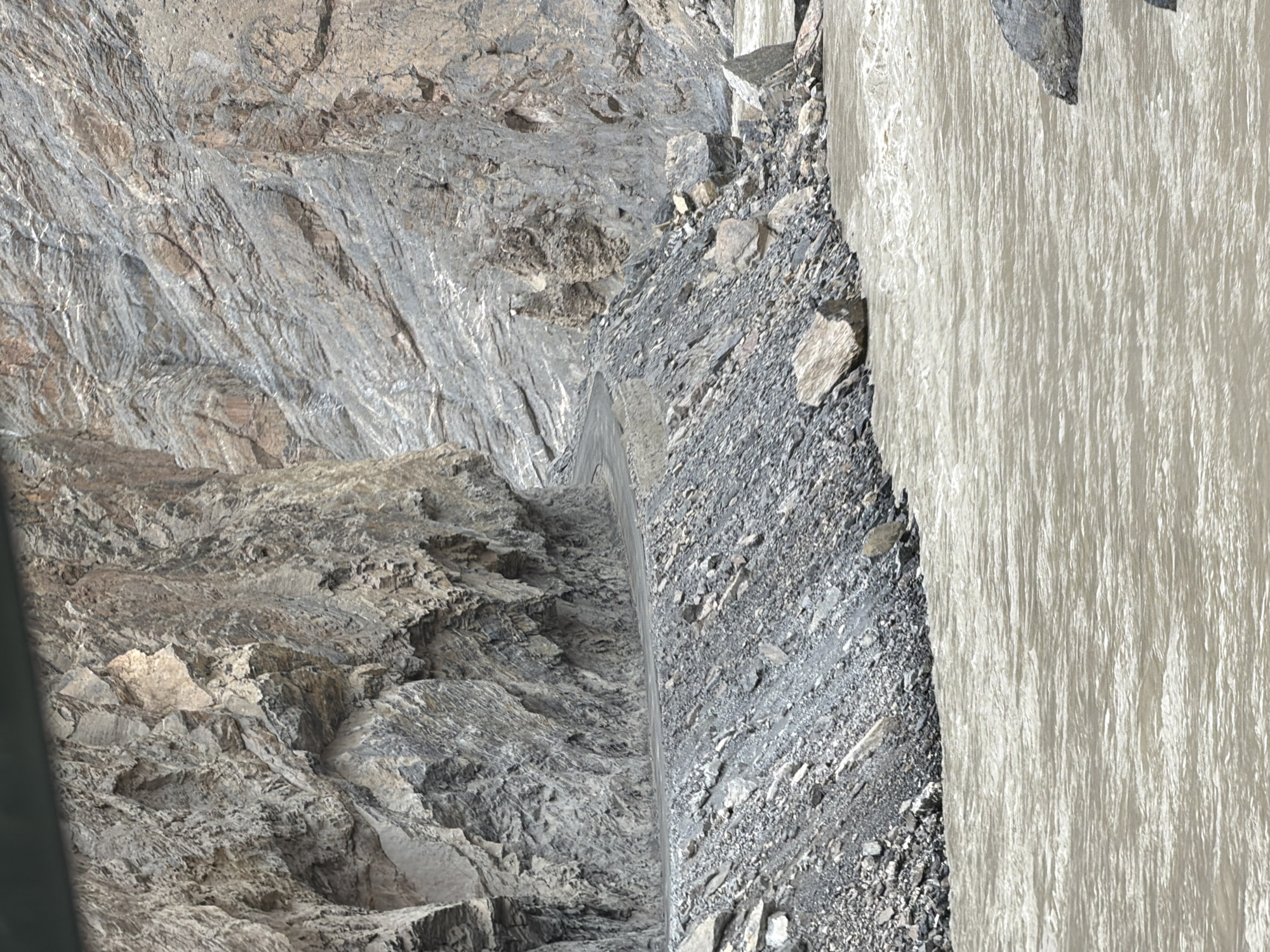 The image depicts a winding road carved into the side of a steep, rocky mountain overlooking a fast-flowing, muddy river. The road appears to be made of gravel or dirt and hugs the cliff face, with loose rocks and scree lining its edge. The mountain itself is composed of layered, grey rock formations, indicating a significant geological history. The river below is wide and turbulent, with visible ripples and white water suggesting a strong current. The environment appears rugged and remote, characteristic of a mountainous region. The image does not contain any people or specific landmarks that would allow for precise location identification within Stongde, India, beyond its general mountainous and riverine landscape.