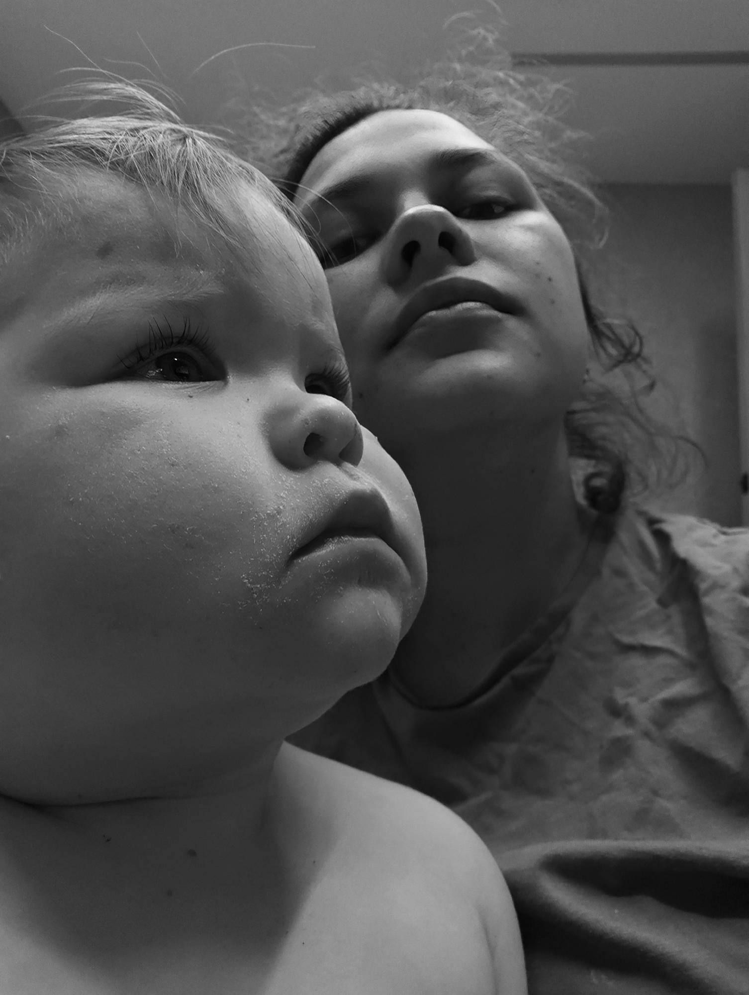 This is a black and white, low-angle, close-up portrait of two individuals, an adult and a child, likely taken indoors in Chaikovsky, Russia. The child is prominently featured in the foreground, positioned on the left side of the frame and in sharp focus. They have light-colored hair, visible eyelashes, and full cheeks, with a thoughtful or slightly solemn expression as they gaze upwards and to the right. Specks of a light-colored substance, possibly food residue, powder, or dried moisture, are visible on their cheek and around their mouth. The child appears to be topless or wearing a light-colored garment that blends with their skin.
Behind and to the right of the child, an adult's face and upper body are partially visible and out of focus. The adult's chin, mouth, nose, and parts of their eyes and forehead can be seen, with their curly, somewhat disheveled hair framing their face. They are also looking upwards, though their expression is indistinct due to the blur and angle. They are wearing a casual, possibly rumpled, light-colored shirt. The background is simple and indistinct, suggesting an intimate, informal indoor setting. The image conveys a quiet, personal moment between the two subjects. No visible text is present.