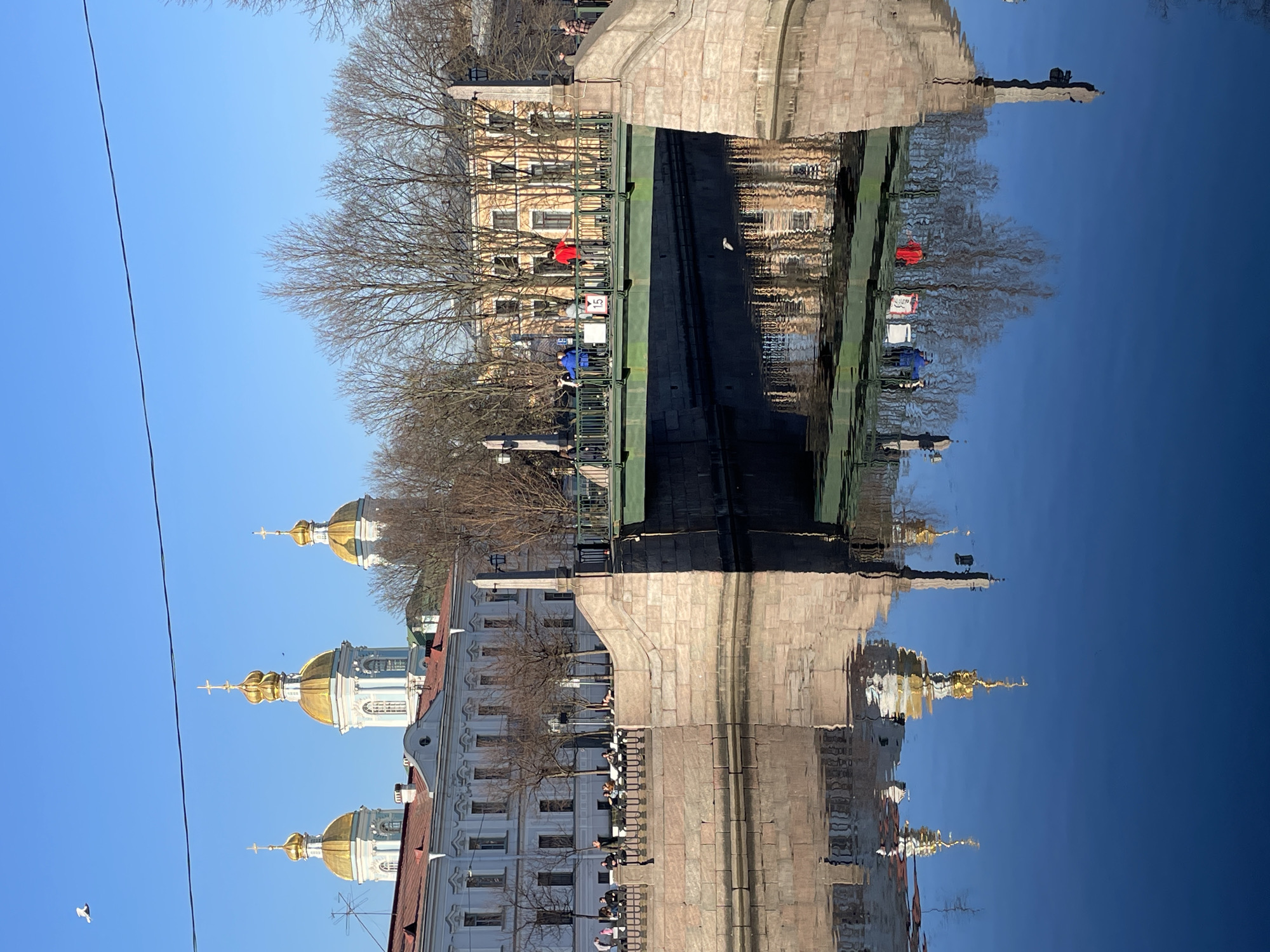 The image depicts a sunny daytime scene along the Kryukov Canal in Saint Petersburg, Russia, featuring the Staro-Nikolsky Bridge. In the background, the Naval Cathedral of St. Nicholas is visible with three prominent golden domes topped with Orthodox crosses, rising above a multi-story white building with a red roof on the left canal bank.
The Staro-Nikolsky Bridge, a green metal structure with decorative railings, spans the canal, supported by stone abutments. On the bridge, at least three individuals are present; one wears a red jacket, another a blue jacket, and a third person is further left. A white sign marked with "15" is affixed to the bridge railing. Bare-branched trees line both sides of the canal and are present behind the bridge. A multi-story yellow building is situated behind the bridge on the right bank.
Along the left canal embankment, adjacent to the white building, approximately five to seven individuals are seated or standing. The calm, dark blue water of the Kryukov Canal reflects the bridge, buildings, cathedral domes, and the clear blue sky with distinct clarity. The sky itself is bright and cloudless, traversed by a single black overhead wire in the upper frame. A white bird is captured in flight in the upper left portion of the sky.