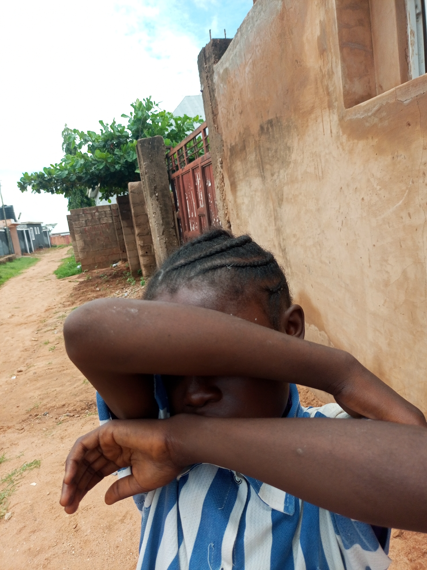A young child is the subject of this media file, with their face hidden by their arms. They are wearing a blue and white striped shirt. The setting appears to be a rural or semi-rural area, with a dirt road leading into the distance. On the right side of the image, there is a tan-colored building with a window opening. Behind the child, a fence made of red metal and brick pillars can be seen, along with a tree with lush green leaves. The sky is visible at the top, with some clouds present, suggesting it might be daytime with partly cloudy weather. The overall mood is somewhat obscured by the child's gesture, which could indicate shyness, sadness, or a desire for privacy. The location is identified as Tempure, Nigeria. There is no visible text in the image.