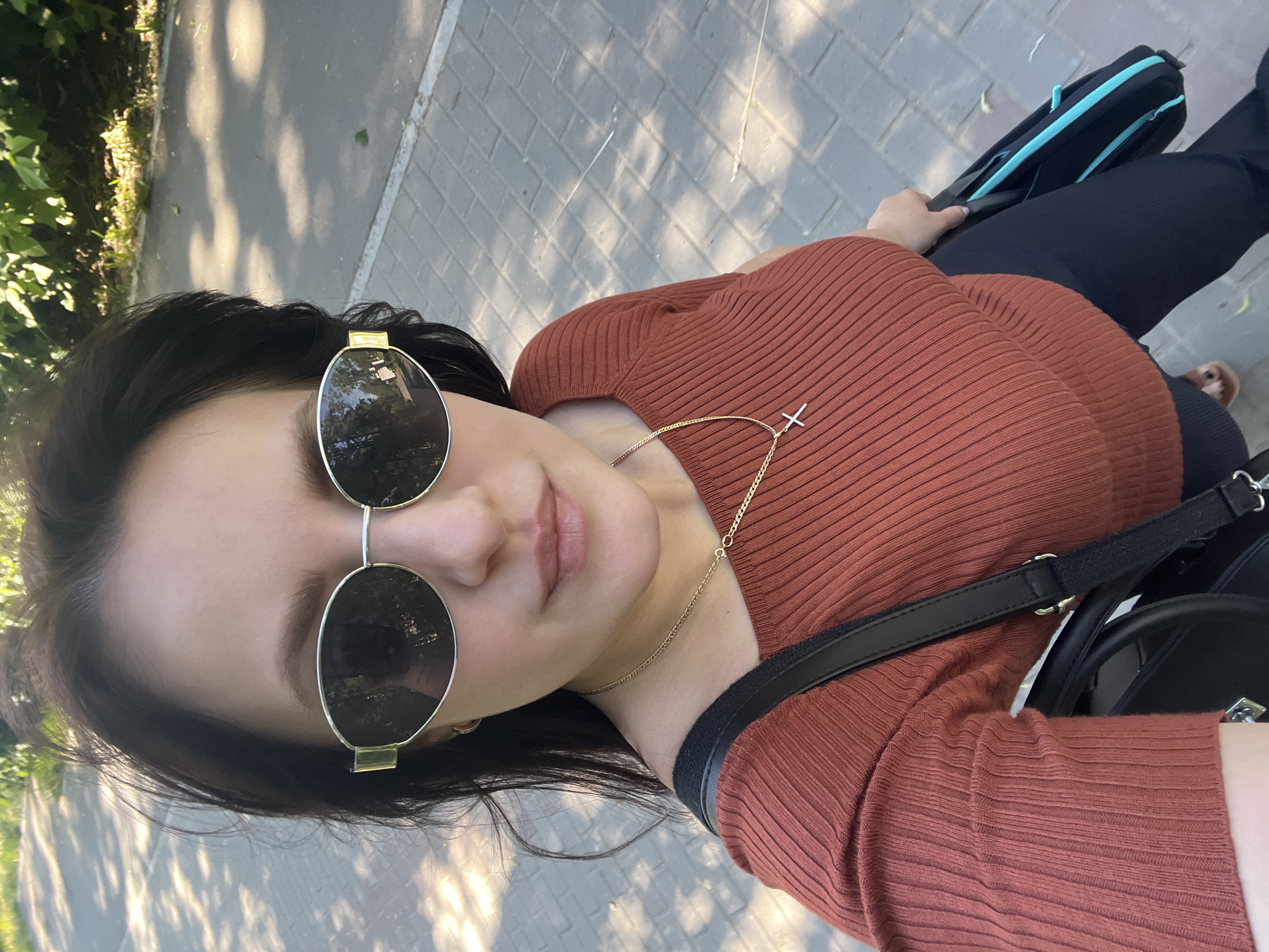 A close-up selfie is taken from a high angle of a woman wearing sunglasses and an orange, ribbed, boat-neck top with a gold chain necklace featuring a cross pendant. She is outdoors on a paved walkway with dappled sunlight and shadows from trees. She is holding a black bag with blue trim. The background shows a sidewalk and green foliage. The image appears to have been taken in Chaikovsky, Russia, given the context of previous images.