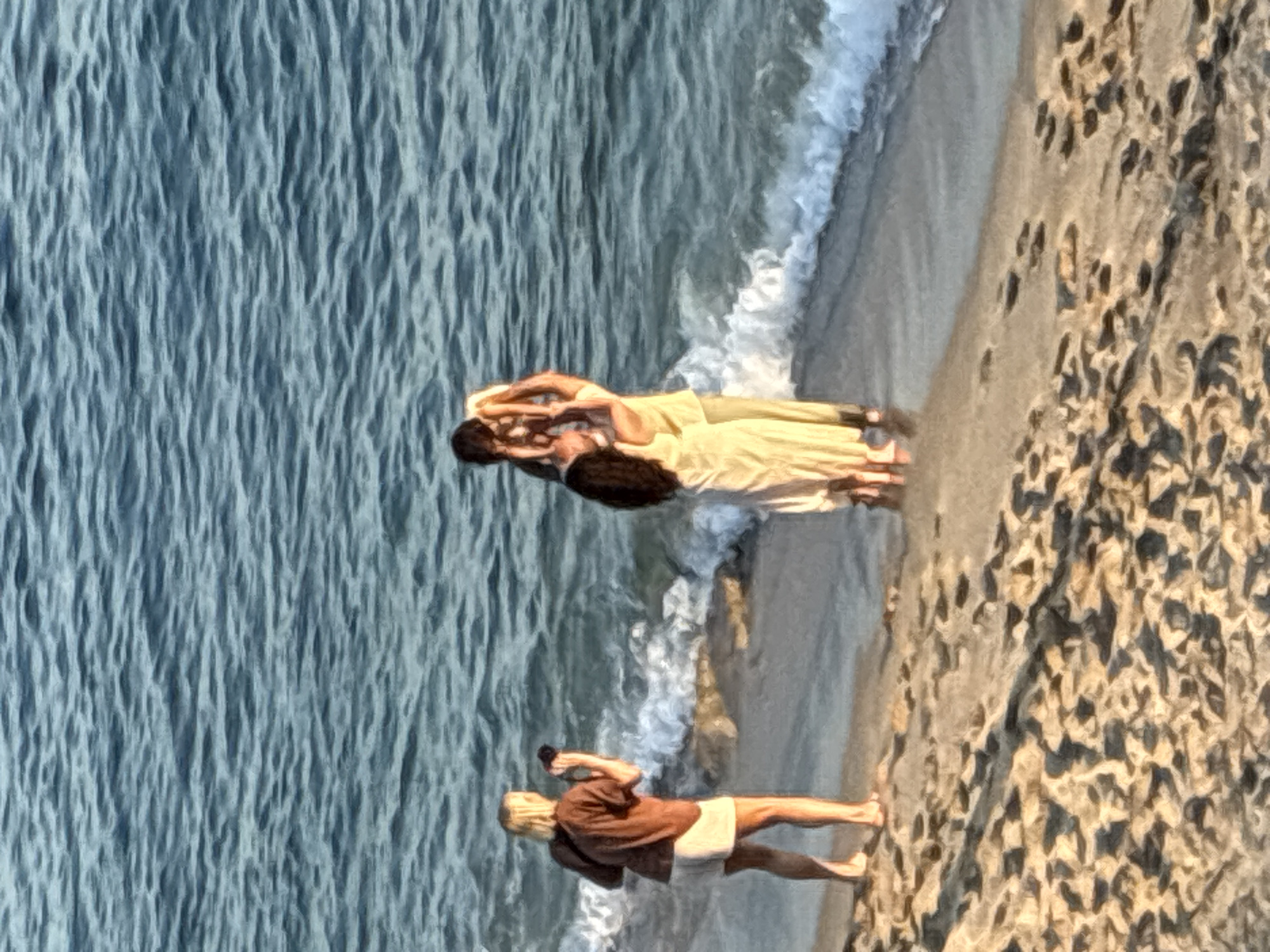 In Alanya, Turkey, on a sandy beach next to the shimmering Mediterranean Sea, three people are captured in a candid moment. The scene unfolds as a woman with blonde hair, wearing a brown t-shirt and white shorts, stands by the water's edge, holding a black camera, seemingly taking a photograph. A couple stands closer to the sea, with a man holding a young child aloft. They are dressed in light-colored clothing, with the woman in a pale yellow dress and the man in a cream-colored outfit. The gentle waves of the sea lap at the shore, creating a tranquil atmosphere. The sand is imprinted with the subtle marks of footprints, suggesting recent activity on the beach.