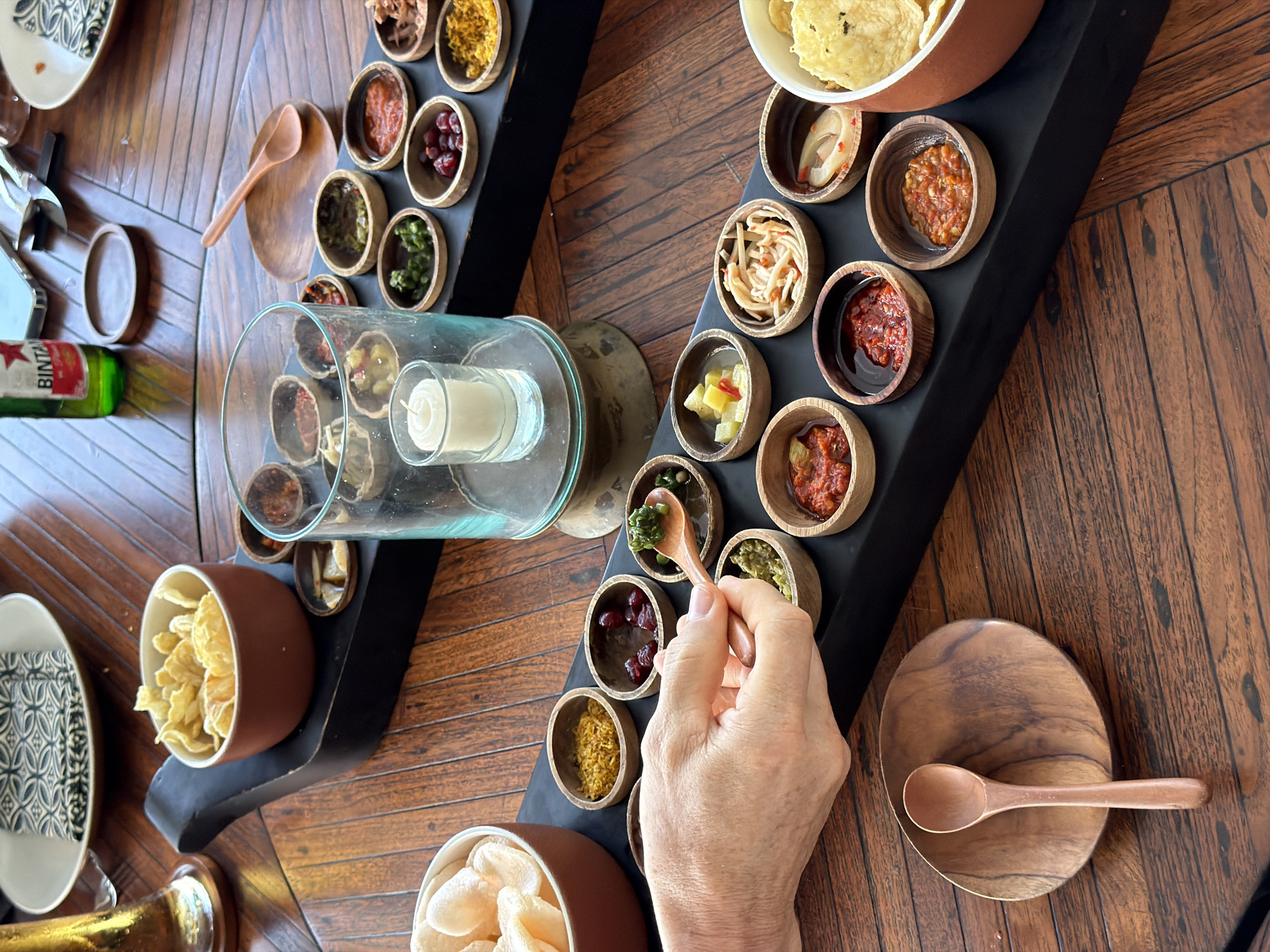 The image captures a dining scene in Pecatu, Indonesia. It features two black serving trays filled with numerous small wooden bowls, each containing different kinds of appetizers or condiments. These are arranged on a wooden table. A cylindrical glass container, holding a lit white candle, sits between the trays. A hand is visible, holding a small wooden spoon, dipping it into one of the small bowls. There are also several bowls of crisps on the table, and a plate with a black and white patterned napkin. An unopened bottle of Bintang beer also lays on the table.
