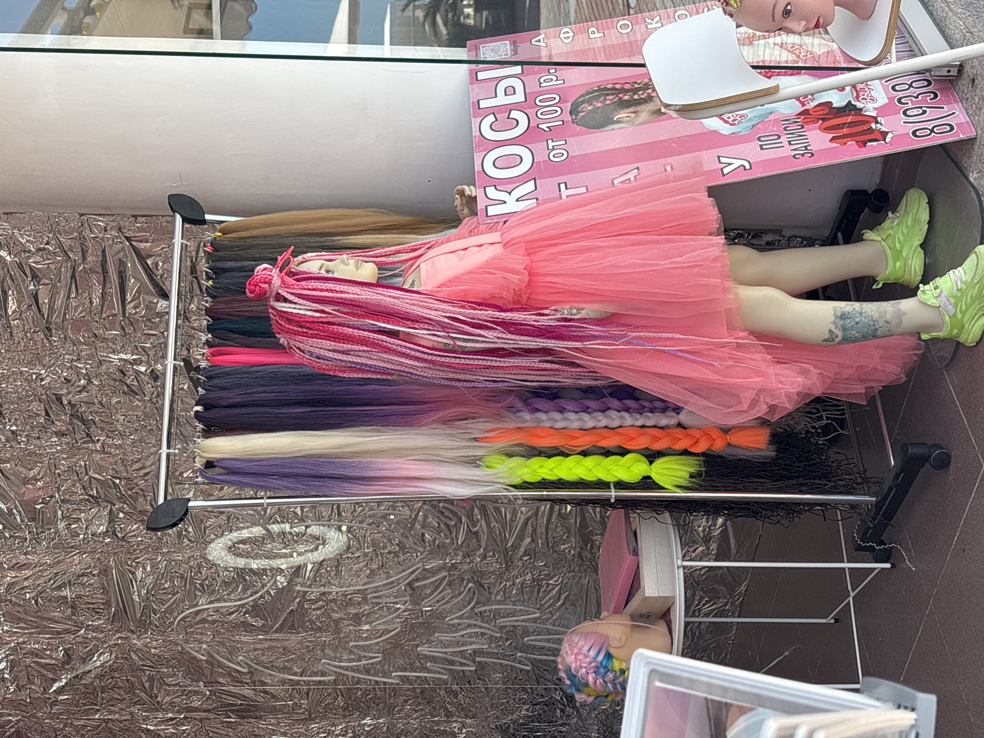 This image captures a vibrant display for hair braiding services and extensions, likely in an outdoor or open-air setting in Sochi, Russia.
The central subject is a blonde mannequin lying on its back, sporting long, vivid pink and white braided hair extensions that extend over its body. It is dressed in a light pink, layered tulle dress and striking neon green chunky sneakers. A notable detail is a blue floral and butterfly tattoo visible on its right leg.
To the left of the mannequin, a metal rack holds an extensive collection of hair extensions arranged in bundles. These come in a diverse palette of colors, including various shades of brown, blonde, purple, bright orange, and neon green, with some featuring ombre effects. The ground beneath this display is covered with crumpled silver foil, adding a reflective texture to the scene.
Behind the mannequin's head, a pink rectangular sign is propped up, serving as an advertisement. The text on the sign is in Russian and clearly states "КОСЫ" (Braids) and "от 100 р." (from 100 rubles), indicating the cost. Another visible phrase is "ПО ЗАПИСИ" (by appointment), suggesting services are booked in advance. A partial phone number "8 (938)..." is also discernible. The sign also features an illustration of a person with braided hair.
In the bottom right corner, a smaller mannequin head with multi-colored braided hair (pink, blue, yellow) is partially visible on a lower display level. The overall impression is a lively and colorful advertisement, showcasing trendy hair styling options during what appears to be daytime.