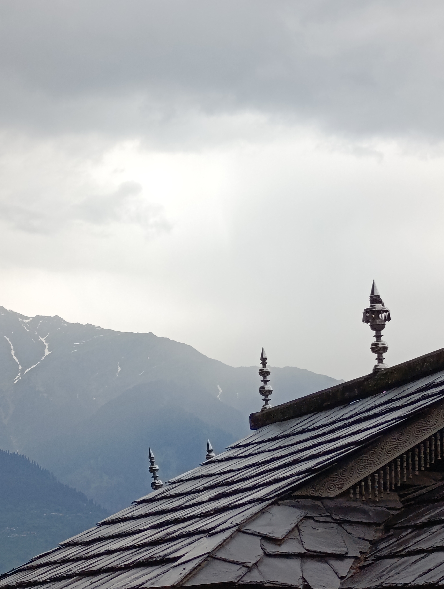 The image captures a close-up perspective of a traditional building's roof set against a mountainous landscape under an overcast sky, likely in Naggar, India. The prominent subject is a dark, multi-layered roof constructed from slate tiles, giving it a rustic and robust appearance. Along the ridge of the roof, several decorative metallic finials, resembling spires with tiered sections, stand out, reflecting some light despite the cloudy conditions. Below the roofline, intricately carved wooden eaves with ornate patterns and hanging elements add to the traditional architectural style.
In the background, a grand mountain range rises, partially enveloped by mist or low-hanging clouds. Patches of snow are visible on the higher peaks and crevices of these mountains, indicating their elevation. Hints of a forested valley or lower slopes can be seen between the foreground structure and the distant mountains.
The sky is entirely covered by dense, grey clouds, suggesting an overcast day, possibly hinting at recent or impending rain. The diffused light creates a soft, somewhat dramatic atmosphere. No specific activity or event is taking place, and no people are visible in the frame. There is no discernible text. The scene beautifully combines local traditional architecture with the natural grandeur of the Himalayan region.