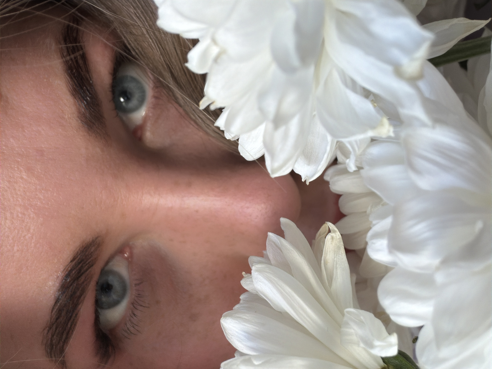 A close-up, tilted shot features a woman's face surrounded by white chrysanthemum flowers. Her left eye is clearly visible, with a prominent eyebrow. The right side of her face is partially obscured by the petals of the flowers. Her skin tone is light, and some freckles are visible. The location is not discernible as Chaikovsky, Russia, or any other specific place.