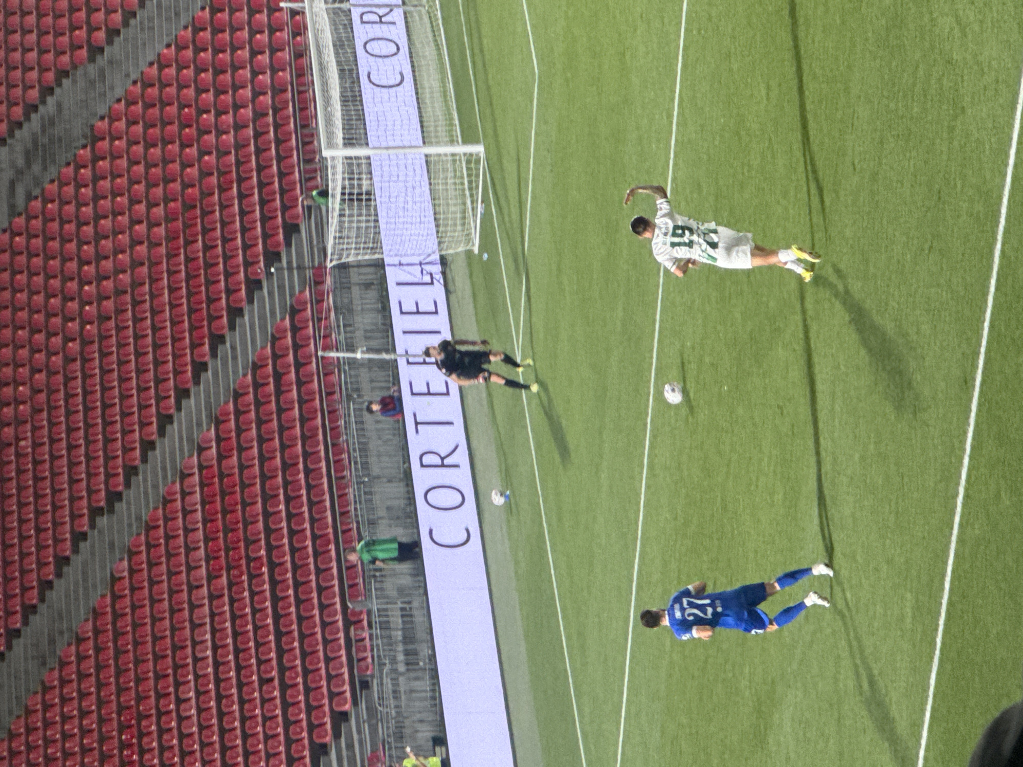 The image depicts a football match being played in a stadium. The stadium has tiered red seating, most of which is empty, and a green artificial turf field. A large white banner with the word "CORTEFIELD" is visible in the background.
In the foreground, on the field, are two football players. One player, wearing a blue jersey with the number 27, is running towards the left side of the frame. Another player, wearing a white and green striped jersey with the number 19, is facing away from the camera, with his right arm raised. A football is visible near this player. Another football is visible further up the field, near a player in a black goalkeeper's uniform.
The image was taken at the Mikheil Meskhi Stadium in Tbilisi, Georgia. The lighting suggests it is daytime, likely late afternoon or early evening given the shadows.