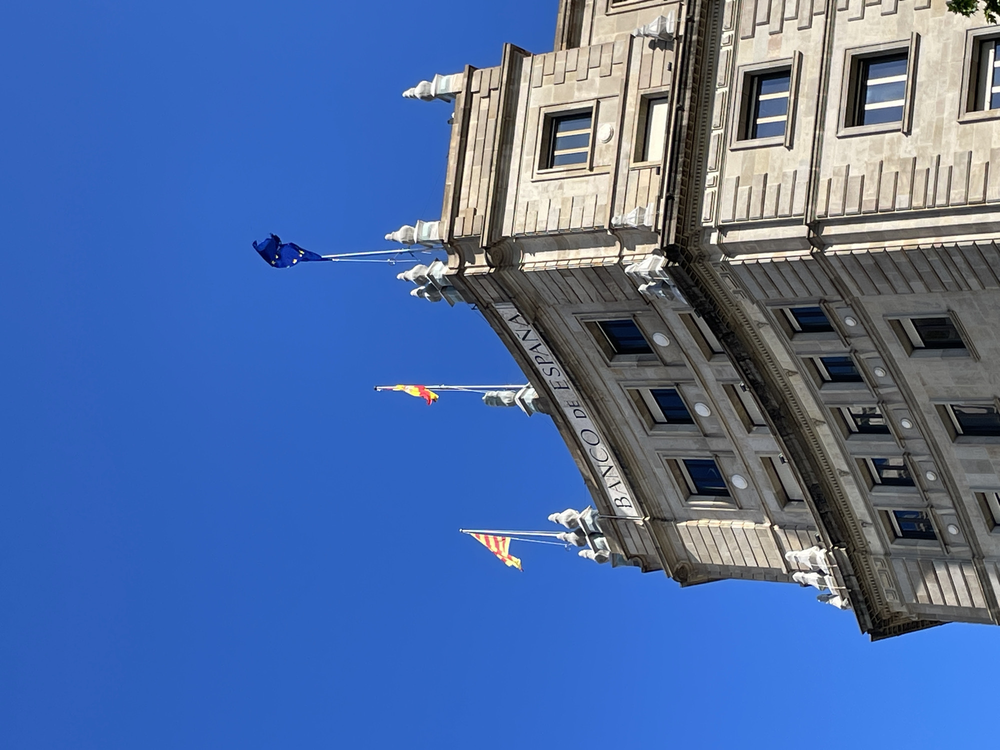 The image shows the Banco de España (Bank of Spain) building from a low angle, likely in Barcelona, Spain. The architectural style is formal and imposing. Three flags are displayed on the building: the European Union flag, the Spanish national flag, and the Catalan flag. The sky is clear and blue.