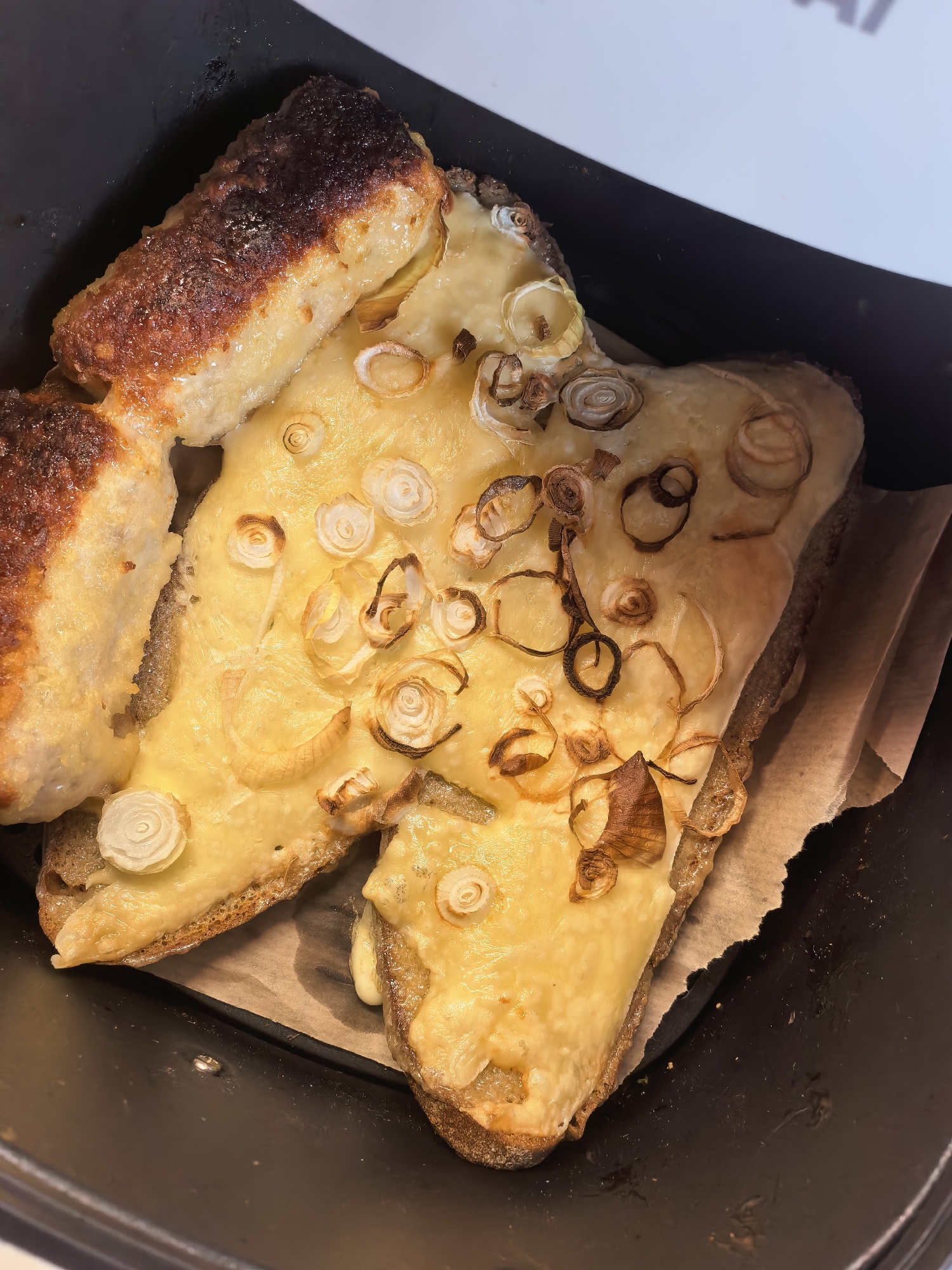 The image shows two slices of open-faced sandwiches topped with melted cheese and thinly sliced, caramelized onions. The sandwiches are placed in a black tray, possibly an air fryer basket, on a piece of parchment paper. One slice of sandwich is partially obscured by another to its left. The bread appears to be dark, possibly rye or whole wheat, with a visible crust. The cheese is a golden yellow and has melted over the bread and onions, creating a slightly gooey texture. Some of the onions are lightly browned and crispy, while others are softer and translucent. The background is blurred, but the overall impression is of a close-up shot of freshly prepared food. There is no information about the location within the city.