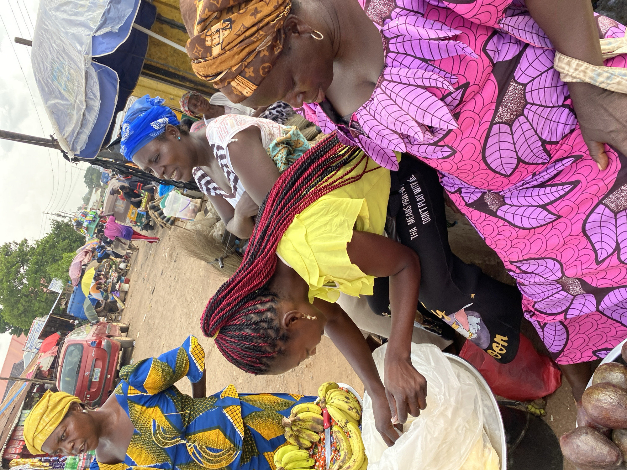 The image depicts an outdoor market scene in Bukuru, Nigeria. Several women are present, some appearing to be vendors and customers. One woman in the foreground wears a yellow headwrap and a blue and yellow patterned dress. To her right, another woman with red and black braided hair wears a bright yellow top and is reaching into a plastic bag. She is standing next to a large bowl filled with bananas and a knife. A third woman, further back, is dressed in a vibrant pink and purple patterned dress and a brown headwrap. She appears to be interacting with someone off-camera. In the background, various market stalls are visible with umbrellas providing shade, and a red car is parked. The ground is dusty and uneven, typical of an outdoor market. The lighting suggests it is daytime, and the sky is overcast. The overall atmosphere appears busy and transactional. Visible text includes "DON'T PLAY WITH ME" and "THA MEANS PRA..." on the black pants of the woman in the pink and purple dress. There is also text on signs in the background.