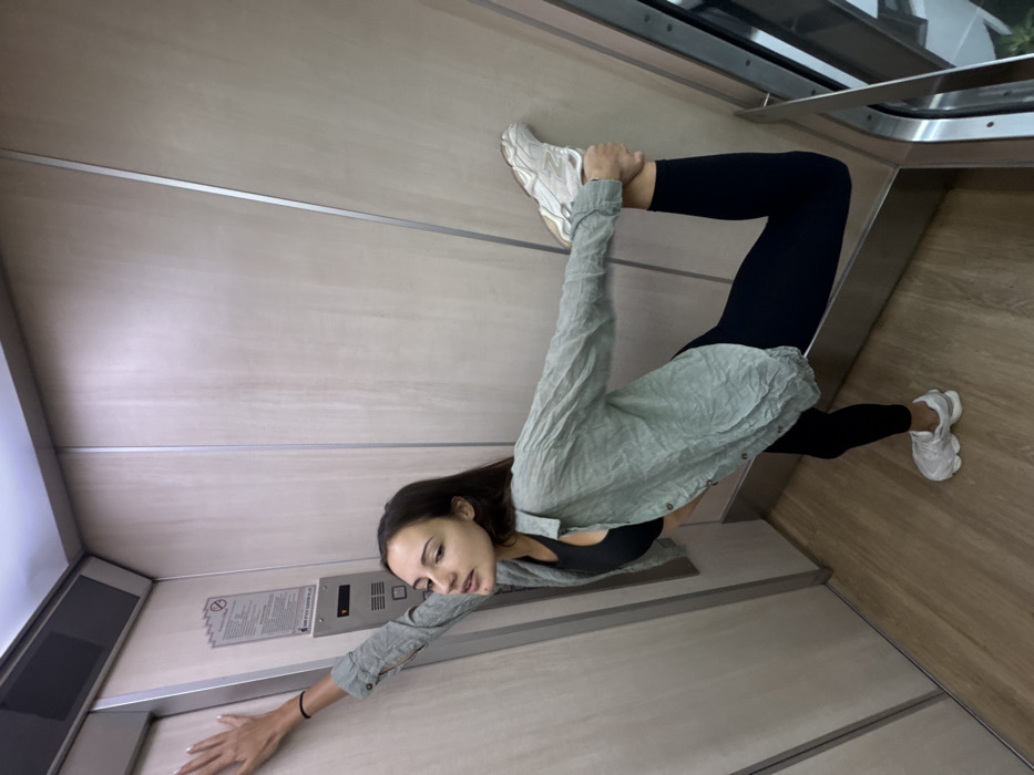 The image displays a young adult female inside an elevator, identified as being in Bangkok, Thailand. The elevator interior consists of light-toned wooden or laminate wall panels accentuated by vertical silver-colored strips, complemented by a matching light wooden floor. On the left wall, an elevator control panel with buttons and a digital display is visible beneath a notice sign featuring text in both Thai and English, and a "No Smoking" symbol. The female subject has long, dark hair and is dressed in a black sports bra, black leggings, a loose-fitting, unbuttoned light green long-sleeve shirt, and white athletic shoes. She is depicted performing a stretching or yoga pose: her left arm is extended upwards with her hand flat against the left wall for balance, while her right hand grasps her right foot, pulling the leg back and up towards her back, bending at the knee. She is balanced on her left leg and looks directly at the viewer with a neutral expression. A partial view through a glass panel on the far right reveals an external urban building.