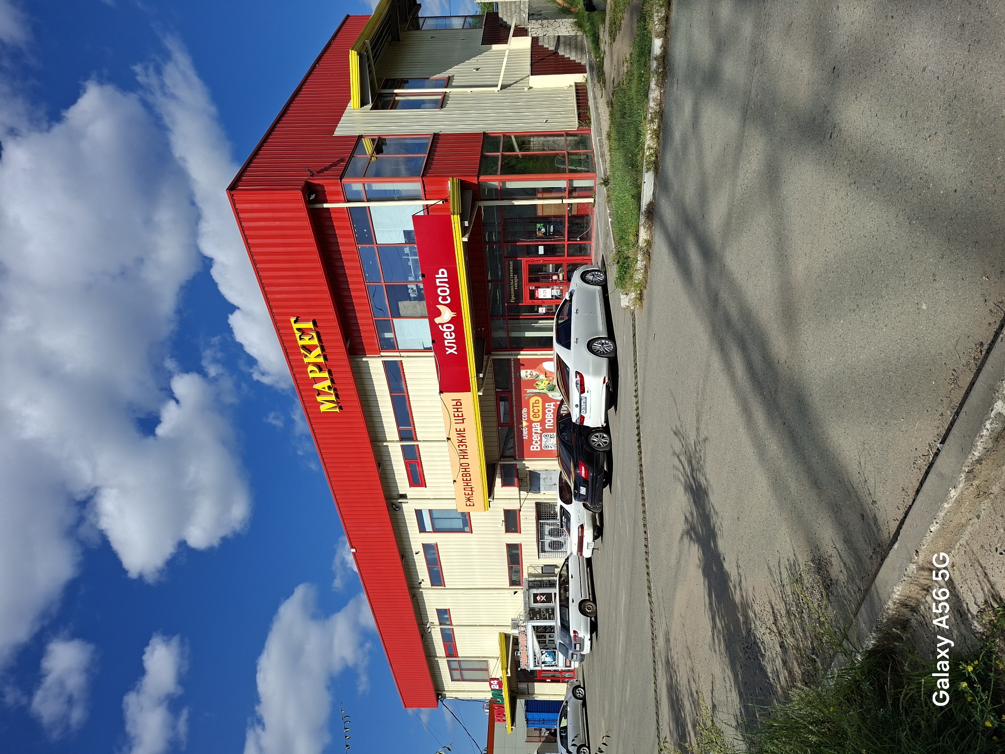 The image shows a "Market" (МАРКЕТ) building with a red facade and yellow accents, located in Chita, Russia. The building has large windows and an entrance with a prominent red awning featuring the text "хлеб соль" (bread salt). Several cars are parked in the lot in front of the building, including a white Toyota Camry, a black Mercedes-Benz, and a white SUV. The sky is blue with scattered white clouds, and sunlight casts shadows on the asphalt. A sign on the building reads "ЕЖЕДНЕВНО НИЗКИЕ ЦЕНЫ" (daily low prices). Another smaller sign visible on the building says "Павлин 24" (Peacock 24). The lower left corner of the image displays "Galaxy A56 5G".
