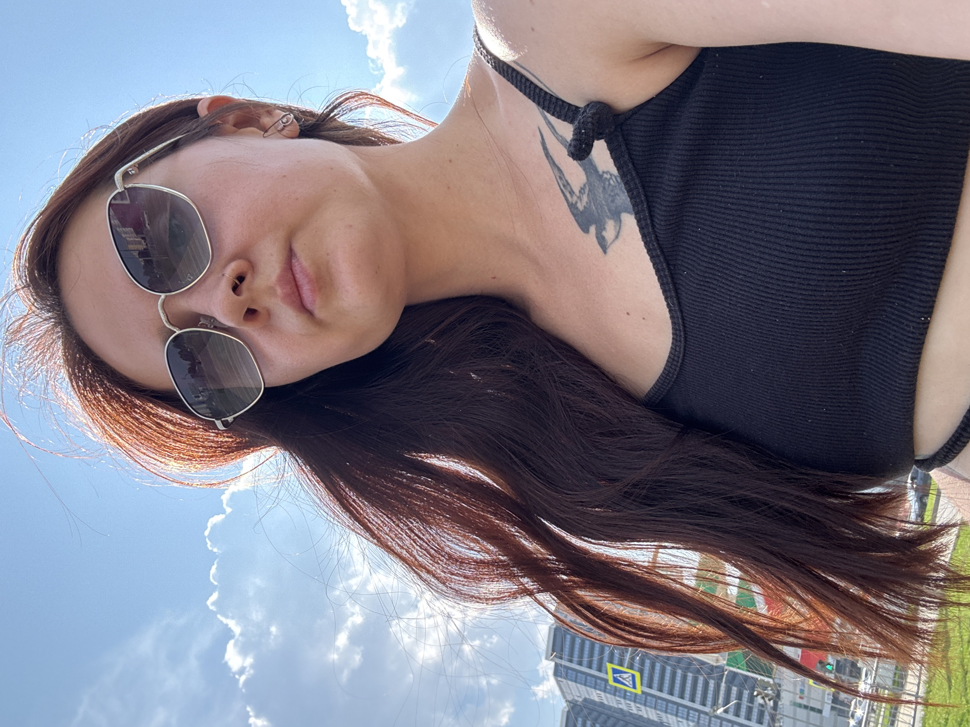 A young woman with long, dark reddish-brown hair and fair skin is pictured outdoors on a sunny day. She is wearing silver-rimmed sunglasses and a black ribbed tank top. A black bird tattoo is visible on her left collarbone. The background shows a bright blue sky with scattered white clouds, and in the distance, the modern architecture of buildings can be seen, along with street signs and traffic, indicating an urban environment. This scene is located in the city of Naberezhnye Chelny, Russia.