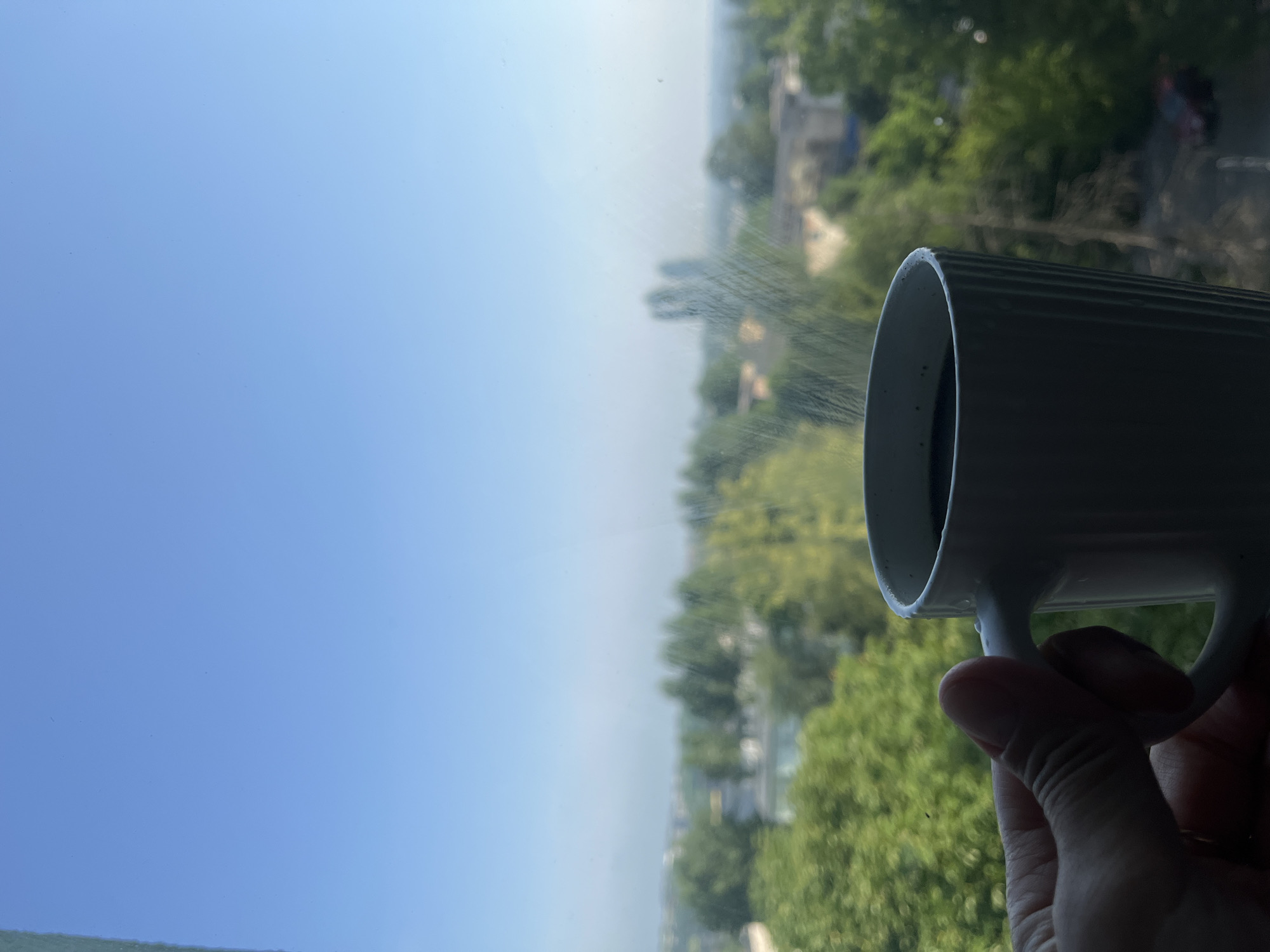 A hand holds a white mug of dark coffee against a backdrop of a hazy city skyline and a bright blue sky. The mug has vertical ridges and sits on a surface that appears to be a windowsill. Faint steam rises from the coffee. The city in the distance is blurred, with greenery from trees and buildings visible. The overall impression is a moment of quiet contemplation with a view.