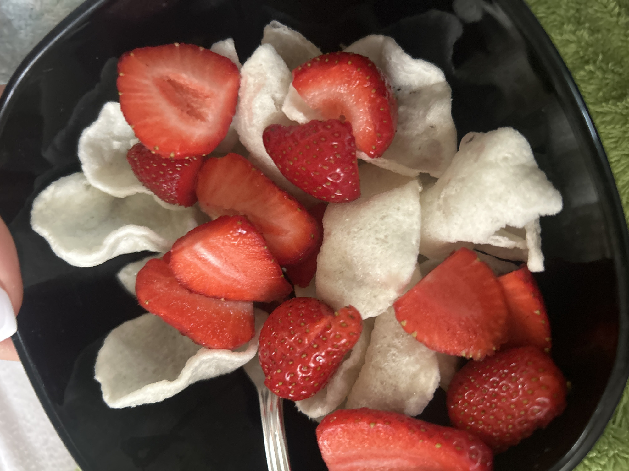 The image shows a black bowl filled with sliced strawberries and white prawn crackers. A silver fork is visible within the bowl. The setting appears to be indoors, with a white surface and a green object in the background. The city and specific location are not discernible from the image.