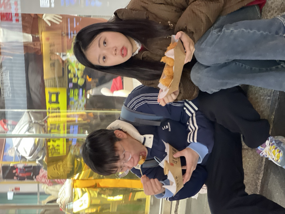 Two East Asian individuals are seated on a grey stone step or ledge in front of a brightly lit storefront, consistent with a commercial street within Yuzhong District, Chongqing, China, during the evening or night.
The male on the left, wearing clear-rimmed glasses, a dark blue hooded jacket with white stripes on the sleeves, and dark pants, is in the act of eating. He uses a wooden skewer to place a piece of fried food from a rectangular cardboard tray into his open mouth. The tray also contains a small cup of sauce. A dark blue backpack is visible on his left shoulder, and he wears multi-colored sneakers.
The female on the right, with long dark hair, wears a brown puffy jacket over a white and red collared top and blue jeans. She holds a similar cardboard tray containing several pieces of fried, golden-brown food items. She looks directly towards the viewer with a neutral, slightly open-mouthed expression.
The background consists of a large glass window, reflecting interior merchandise such as shoes and mannequin parts, as well as exterior neon or illuminated Chinese characters and other commercial signage, indicating a bustling urban commercial area.
