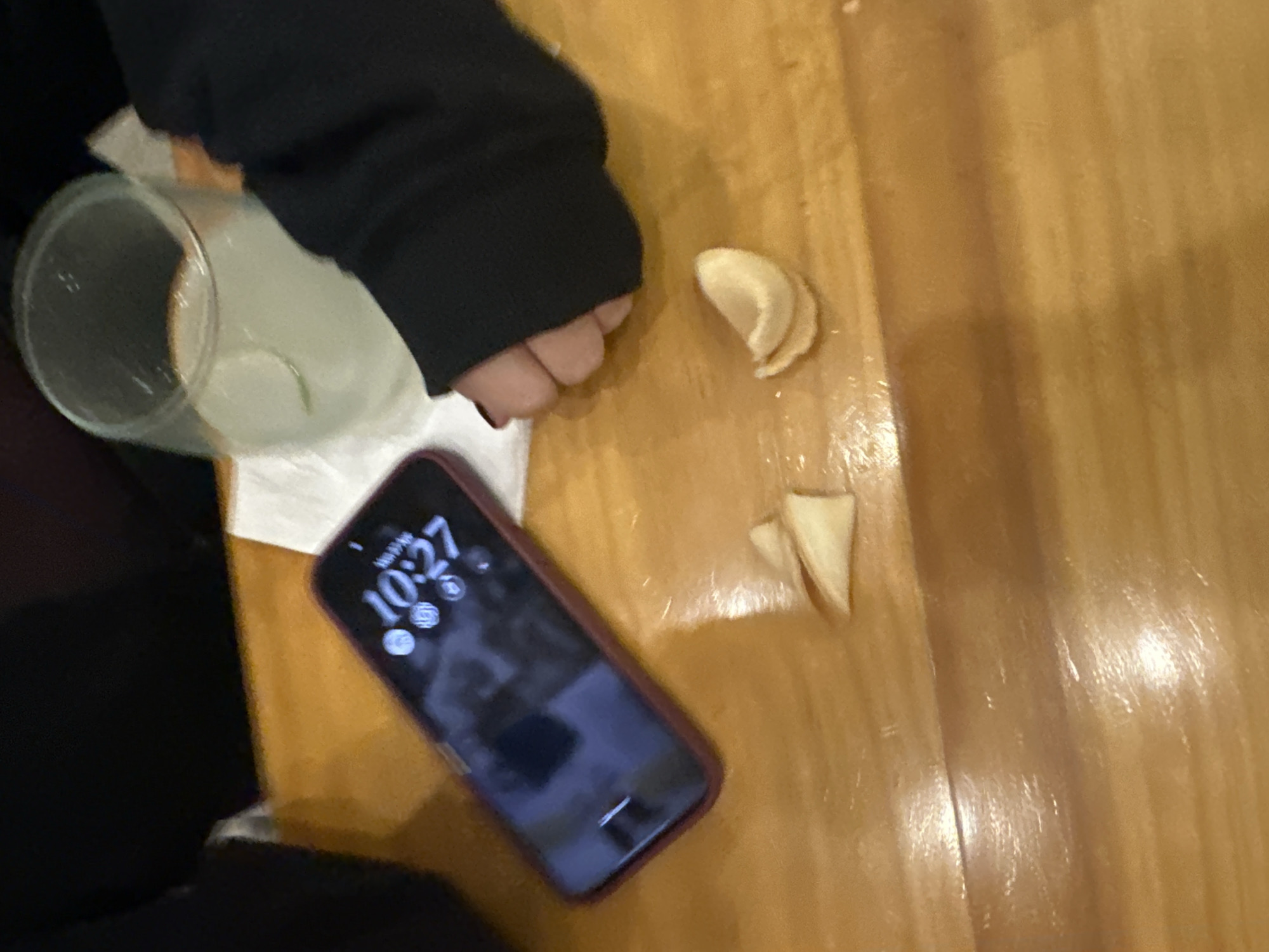 The image captures a slightly blurry, high-angle close-up of a casual indoor scene, likely at a table with a glossy light-brown wooden surface.
A person's left arm, wearing a black long-sleeved top, extends into the upper left of the frame. Their hand is partially visible near a clear glass containing a light green liquid, possibly a limeade, with a slice of lime inside. The glass rests on a crumpled white napkin.
Scattered on the wooden surface near the person's hand are two pieces of a light-colored, curved, and brittle food item, resembling remnants of fortune cookies or fried wonton wrappers.
A smartphone, encased in a dark red or maroon protective cover, lies on the table in the lower central part of the image. Its screen is illuminated, clearly displaying the time "10:27" and "Mon 27," along with various app icons and a dark background.
The wooden surface exhibits visible grain and reflects light, suggesting a polished finish. The scene portrays a moment of relaxation or dining, with a drink and a phone readily accessible. Given the context of Mexico City, Mexico, the drink could be a local limonada (limeade), fitting the casual atmosphere.