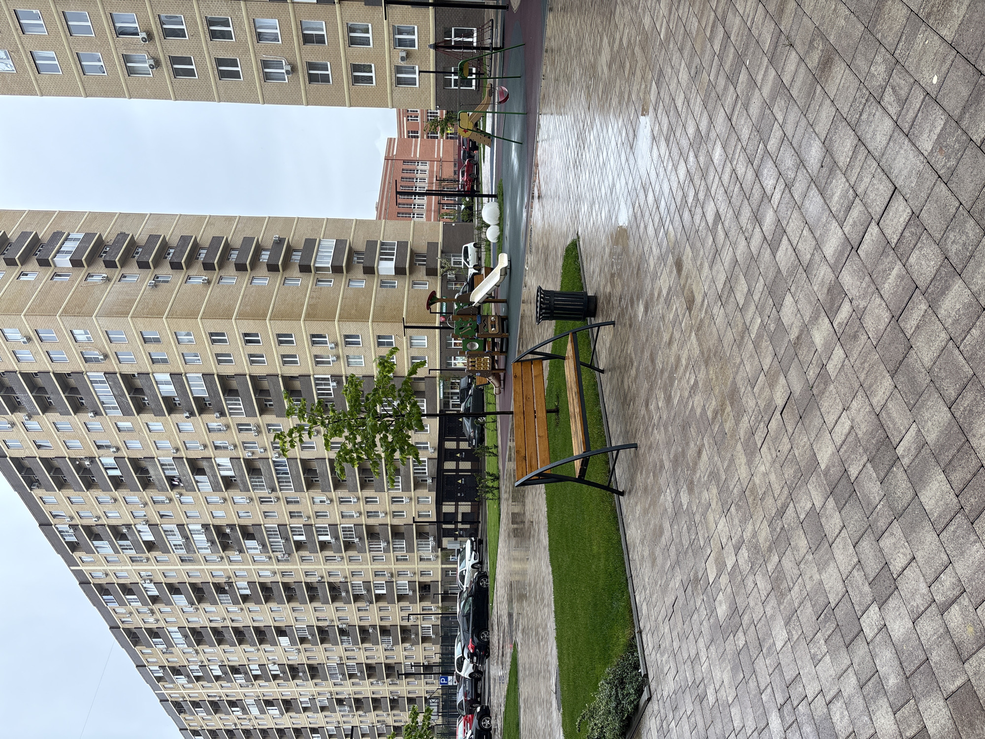 The image captures a quiet, modern residential courtyard or street scene under an overcast sky. The photograph appears to be taken after rain, as the extensive paved area in the foreground and stretching into the mid-ground reflects light from puddles and wet surfaces. This paving consists of interlocking rectangular and square tiles in shades of grey and brown.
On the left side of the frame, a tall, multi-story residential building rises, constructed from light beige or tan brick with numerous windows and recessed balconies that create a textured facade. Several air conditioning units are mounted below windows. A strip of green grass runs parallel to the building and the paved area, where a small tree with green foliage and some low bushes are planted. A wooden bench with a dark metal frame and a dark green trash can are situated on this grassy strip.
Behind the bench and grass, a parking area is visible, with several cars (including white, black, and red vehicles) parked in a row. A blue ground marking with a white "P" signifies parking. In the background, beyond the parking and between the main buildings, a colorful children's playground with slides and climbing structures can be seen. Another tall, multi-story building, similar in style to the one on the left but appearing slightly darker, occupies the background, along with a reddish-brown brick building.
The sky is a uniform light grey, indicating heavy cloud cover. No people are visible in the scene, suggesting a calm and unoccupied moment in the residential area. The visible text is a white "P" on a blue ground sign in the parking area.