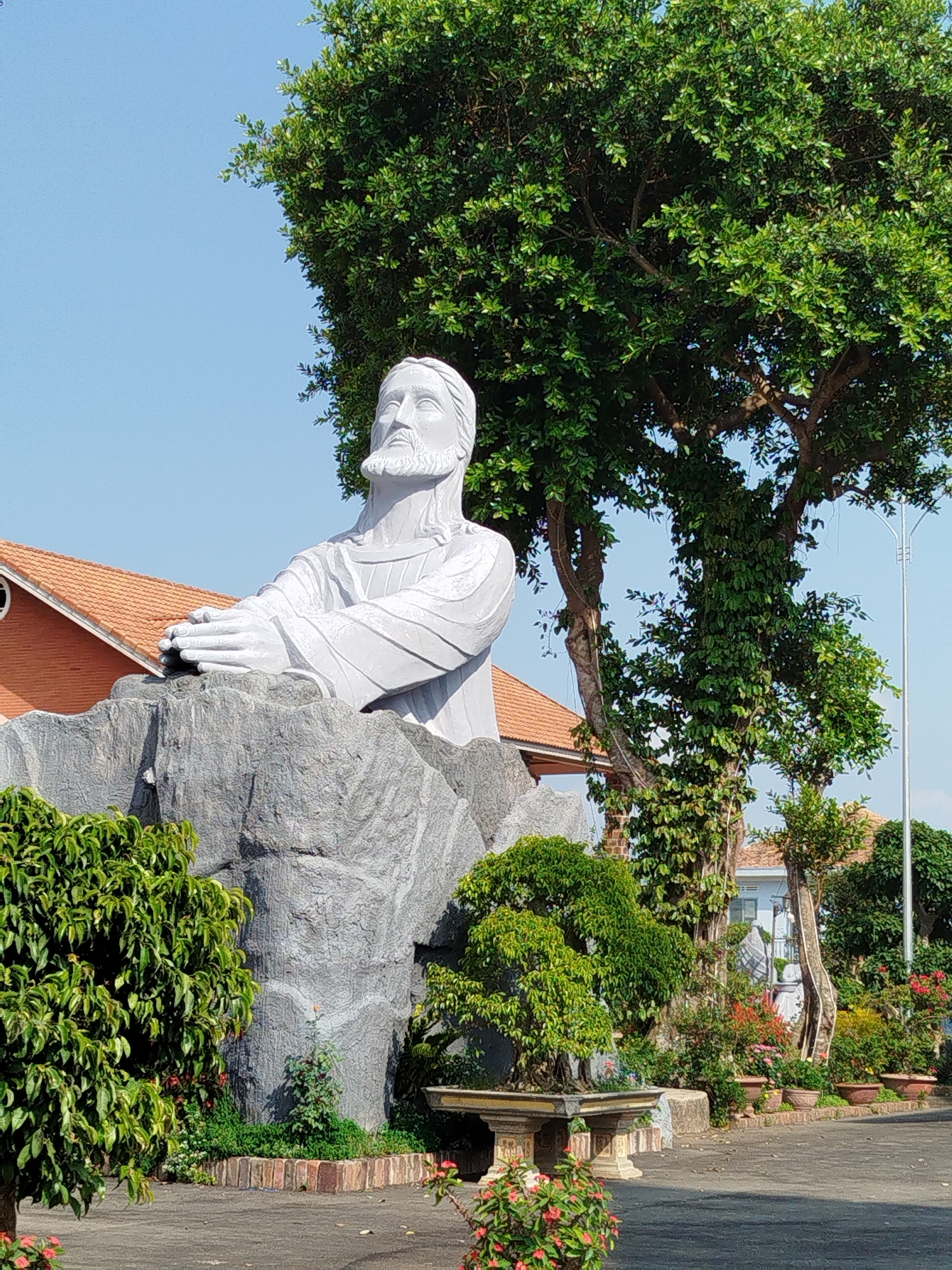 A daytime shot features a gray statue of Jesus in what appears to be a garden setting in Diom A, Vietnam. The statue, which is seated atop a gray, boulder-like base, shows Jesus with his hands clasped in front of him. The statue is white and has a beard and long hair.
Behind the statue is a large, green tree, obscuring much of the blue sky. In the background, sections of buildings with orange-tiled roofs are visible, alongside greenery and additional plants arranged in pots. The foreground consists of paved stone, with additional shrubbery and flowering plants. A small stone bench is also visible amidst the vegetation. The overall setting suggests a peaceful, well-maintained garden or religious ground.