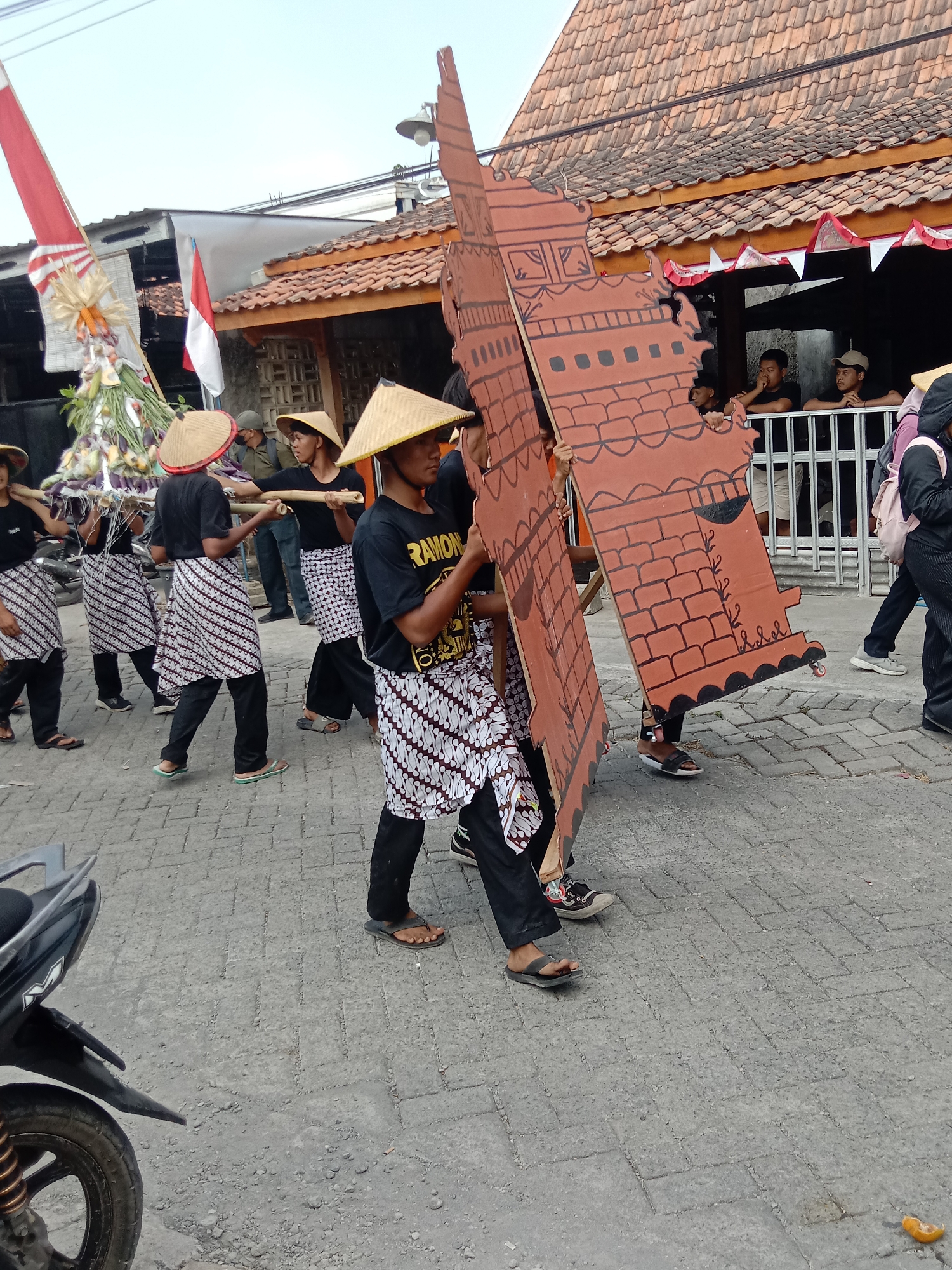 The image depicts a street scene in Wirosari, Indonesia, likely during a procession or cultural event. In the foreground, several individuals are walking, carrying large, flat, cutout cardboard structures painted to resemble brick buildings. These individuals are wearing conical straw hats, dark t-shirts, and patterned sarongs or skirts over dark pants. Some are also wearing flip-flops.
To the left, a tall, conical structure adorned with fruits and vegetables, possibly an offering or part of the procession, is visible. A flag, red and white, is also prominently displayed on a pole. In the background, a building with a terracotta tiled roof and an open facade is present. Behind a metal railing, several onlookers are gathered, some leaning on the railing. A motorcycle is partially visible in the lower left corner of the frame. The ground is paved with interlocking concrete blocks. The sky is overcast.