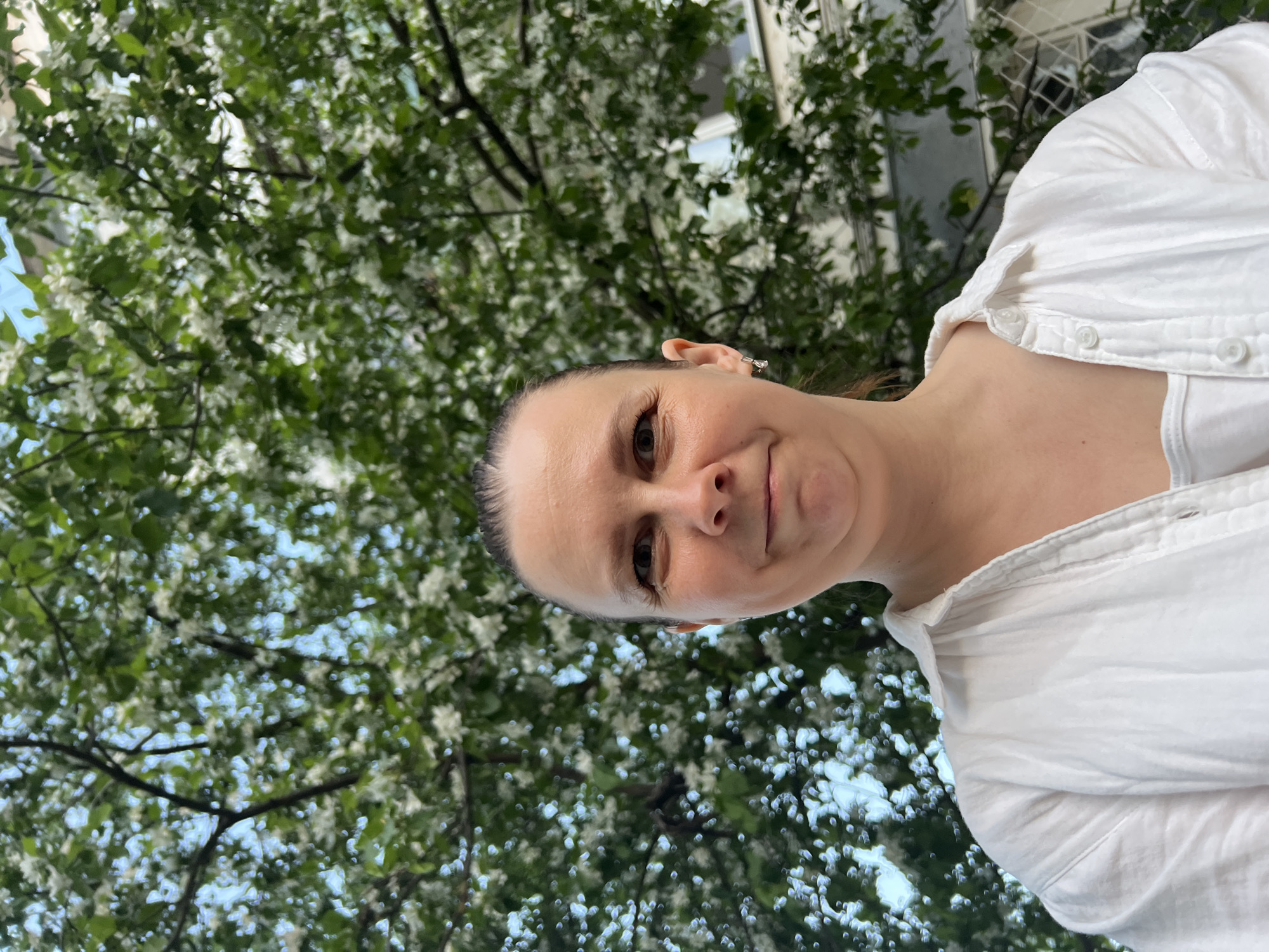 A woman wearing a white collared shirt is facing the camera. The woman is centered. Behind her is a tree with white blossoms. In the background, a building can be seen, possibly in Chaikovsky, Russia.