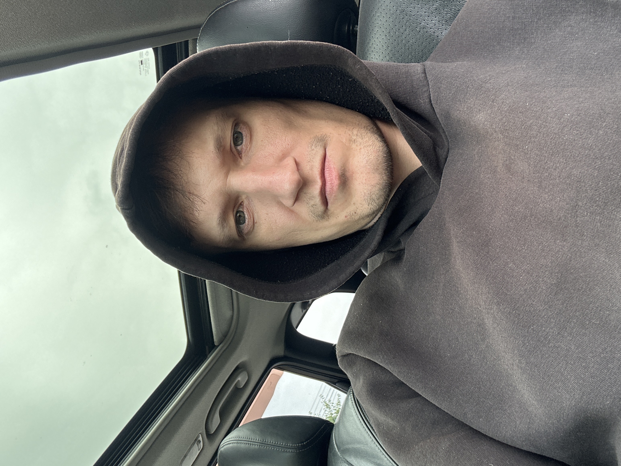 A medium shot selfie captures a man in his late 20s or early 30s, wearing a dark brown hooded sweatshirt, from the chest up. He is seated inside a vehicle, evidenced by the black leather seats and the open sunroof revealing a cloudy sky. The man has light skin, brown hair that is slightly messy, and light-colored eyes. His facial expression is neutral. The interior of the car is dark, and the lighting is subdued. The background shows the car's interior, including the headrest of the passenger seat. No specific location in Makiv, Ukraine, is identifiable from the image.
