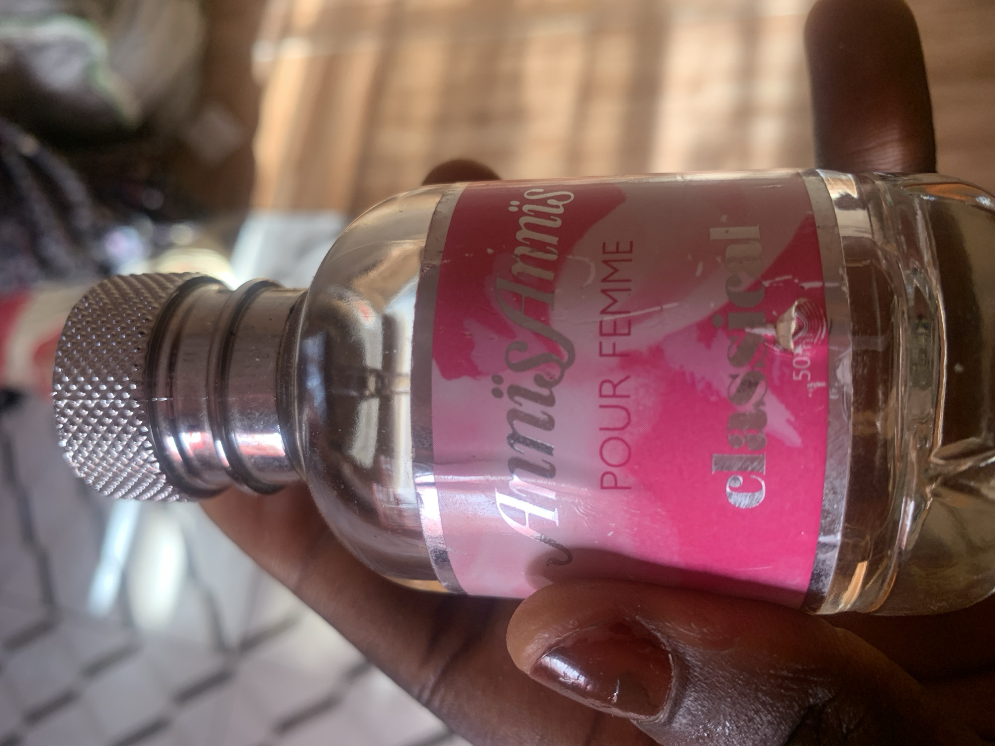A close-up shot shows a clear glass bottle of perfume, labeled "AnniAnnis POUR FEMME Classical 50ml". The bottle has a textured silver cap and is held by a person's hand with dark skin and polished fingernails. The background is blurred, showing a warm-toned, possibly wooden surface, suggesting an indoor setting. The lighting appears to be natural, casting soft shadows and highlights on the bottle and hand, indicating it might be daytime. The focus is on the product, with the hand acting as a frame to present the perfume.