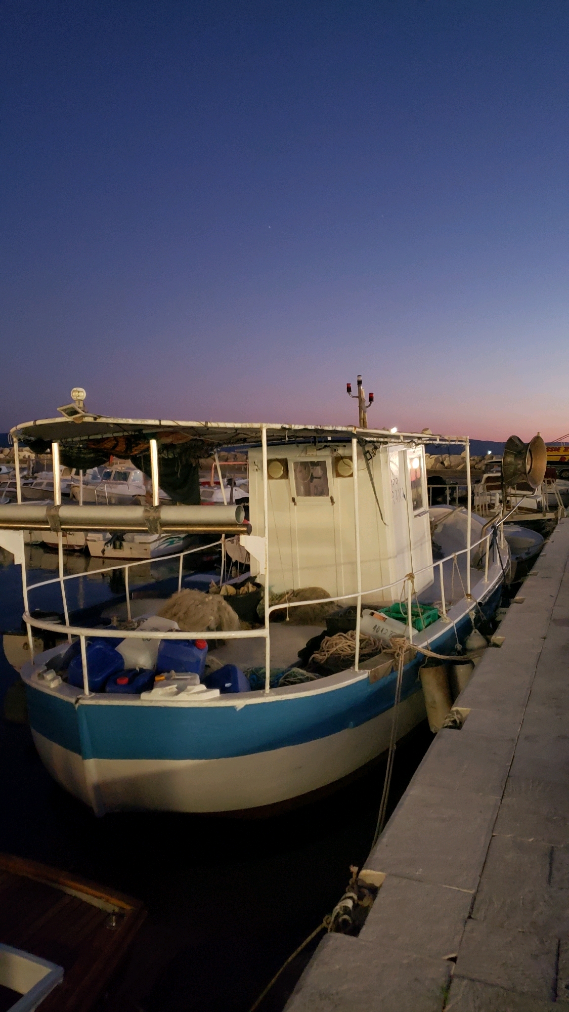 The image depicts a fishing boat moored at a dock in the city of Općina Tučepi, Croatia. The sky displays a twilight gradient, transitioning from deep blue overhead to lighter hues near the horizon, with a few faint stars visible. The boat, primarily white with a blue waterline, is equipped with a covered deck, netting, ropes, and several blue containers. It is tied to a concrete dock, which occupies the right side of the frame. Other boats are visible docked in the background, suggesting a harbor or marina. The lighting indicates it is dusk or early dawn. There are no people visible in the image.